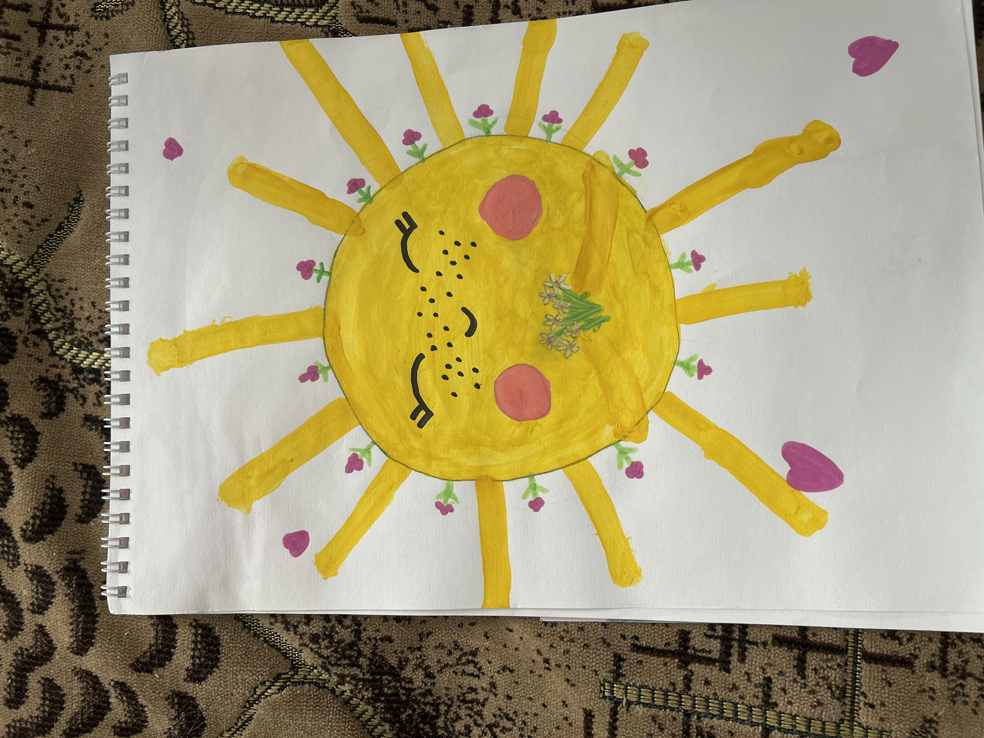 A child's drawing depicts a yellow sun with a smiling face. The sun has closed eyes, pink cheeks, and freckles. Yellow rays radiate outwards from the central circle, and small purple flowers are drawn around the perimeter of the sun. There are a few purple heart shapes floating in the white space surrounding the sun. A small bouquet of white flowers is painted near the sun's cheek. The drawing is on a sheet of paper with a spiral binding, which is resting on a patterned rug. There is no indication of the city of Uray, Russia.