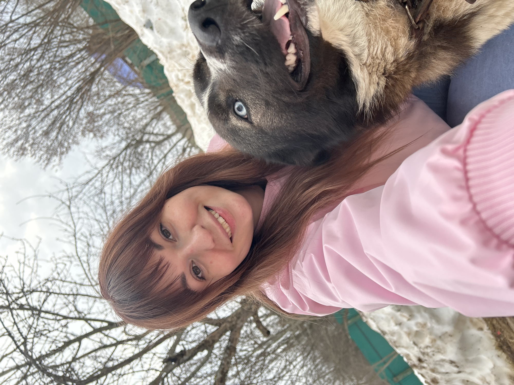 A close-up selfie-style photograph depicts a female subject and a dog outdoors in the city of Фоки, Russia. The woman, positioned on the left, has reddish-brown hair with bangs and brown eyes, smiling directly at the camera with her teeth visible. She is wearing a light pink jacket. To her right, a large dog with black and light brown fur features a prominent blue eye. Its mouth is open, revealing its tongue and teeth, suggesting a panting or joyful expression. A metal chain collar is visible around the dog's neck. The background indicates a winter or early spring environment with snow covering the ground, including a sloped bank on the right. Bare deciduous trees are visible against an overcast sky, along with a section of a teal-green fence behind the snow.