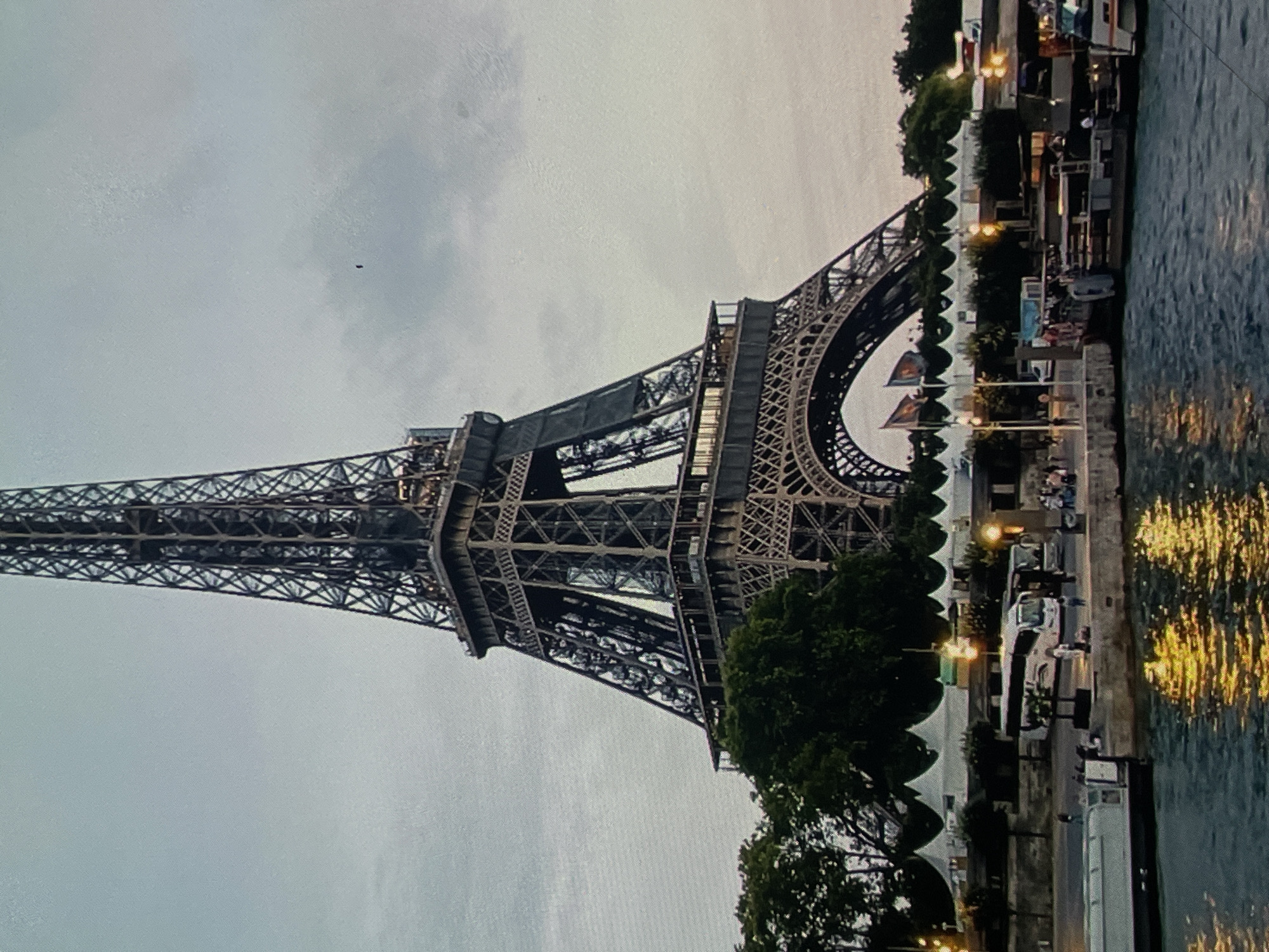 The image shows the Eiffel Tower in Paris, France. The tower is a large iron structure with intricate latticework. In the foreground, there is a body of water, likely the Seine River, with boats docked along the bank. There are also people walking along the riverside promenade, as well as several white tents set up, possibly for an event or market. The sky is overcast, suggesting it might be evening or a cloudy day. The reflection of lights can be seen on the water. There is no indication of Harmanli, Bulgaria in this image.