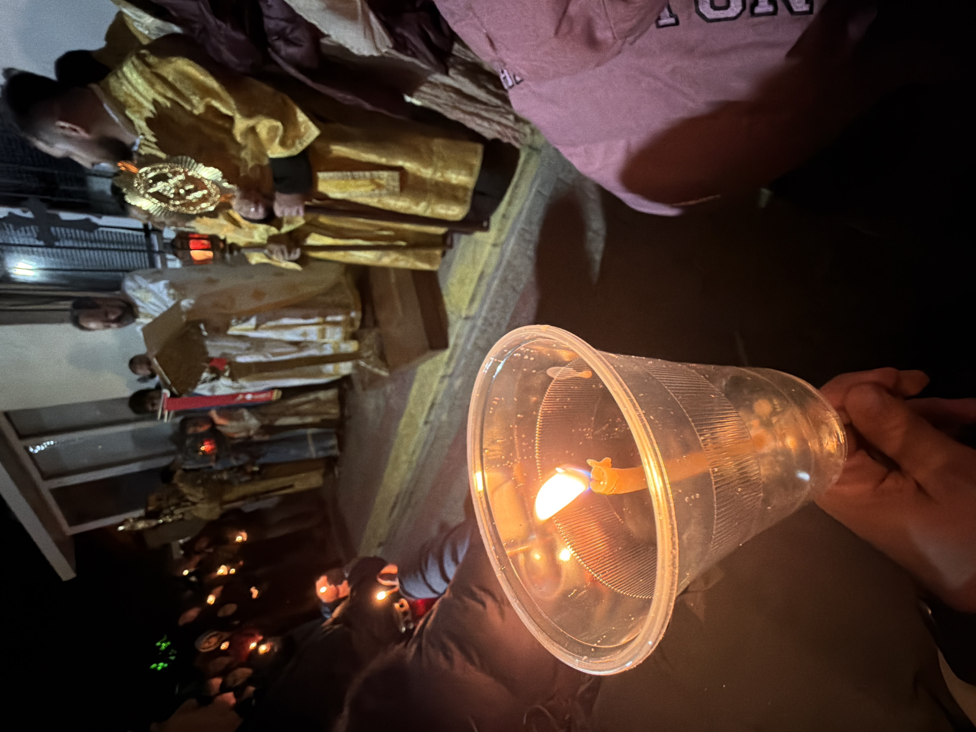 A procession is in progress, illuminated by numerous candles held by participants, some contained within protective plastic cups. The lead participants appear to be religious figures, possibly Orthodox Christian clergy, adorned in gold and white vestments, some carrying ornate religious symbols. The setting is urban, potentially Varna, Bulgaria, given the context. The scene is nighttime, with a focus on the light emanating from the candles. The foreground features a candle in a clear plastic cup, held by a person whose hand is visible. The background shows a line of people also holding candles, with the religious figures positioned further up in the procession. The environment appears to be a paved area, possibly a street or sidewalk, beside a building.