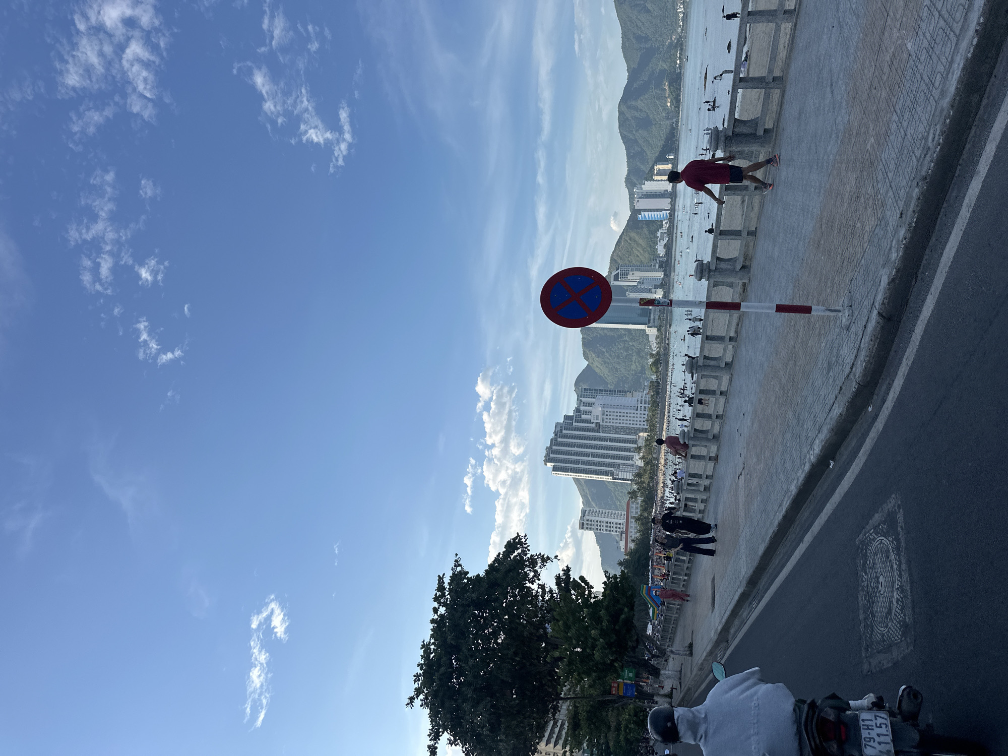 The image depicts a scene on the beachfront promenade of Nha Trang, Vietnam. In the foreground, a person is riding a motorcycle on an asphalt road, with a license plate visible. To the right, a paved walkway runs alongside a concrete railing overlooking a beach. A red and white striped pole stands near a "no parking" sign. Several people are walking or standing on the promenade. Beyond the railing, the beach is populated with many individuals, and the ocean is visible with a few boats. In the background, tall buildings and lush green mountains form the city's skyline. The sky is blue with scattered white clouds.