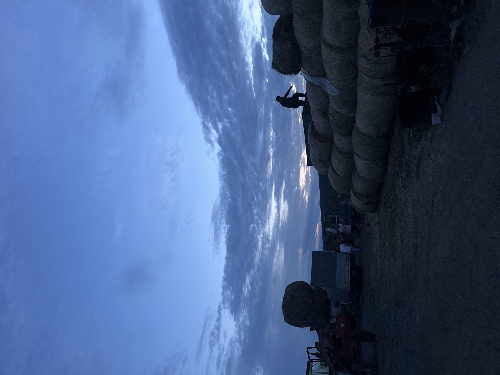 The image, taken in Chubra, Bulgaria, captures a rural farm scene at dusk. The sky dominates the left side of the frame, showcasing deep blue hues with patterned clouds, and a brighter patch of sky on the horizon suggesting the last vestiges of daylight.
On the right side, a substantial stack of round hay bales runs vertically, appearing as a dark, textured wall. Silhouetted against the evening sky, a person stands atop or alongside these hay bales, with arms outstretched or in motion, suggesting activity related to the bales.
Further to the right and towards the bottom, various pieces of farm equipment are visible in shadow, including a red and black tractor, and a trailer or truck with a grey cover. Several other indistinct figures, also in silhouette, can be seen near the machinery, indicating the presence of other people on the farm. The ground beneath is unpaved, appearing to be dirt or gravel. The distant landscape on the far right, possibly hills or mountains, forms a dark silhouette against the horizon. The overall atmosphere is tranquil, characteristic of a farm winding down at the end of the day. No visible text is present in the image.