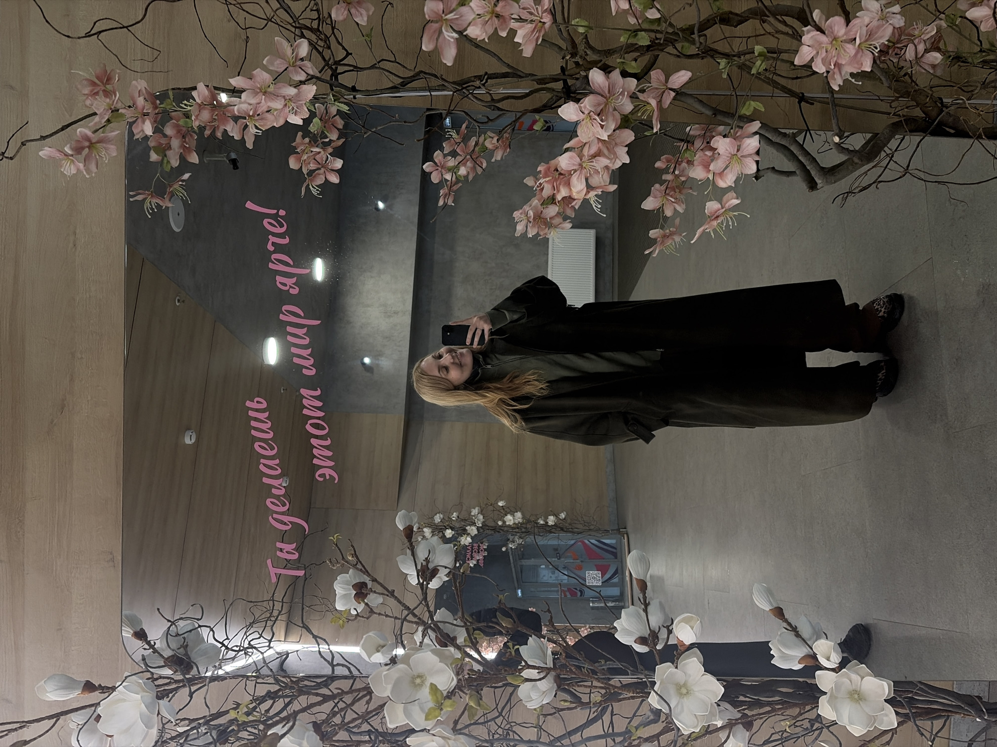 The image depicts a woman taking a selfie in front of a mirror in Izhevsk, Russia. The mirror is decorated with artificial cherry blossoms and magnolia branches. Pink lettering is visible on the mirror in a vertical orientation. The woman is wearing a dark green coat and holding a cell phone. The background shows a public space with grey flooring and various architectural details, including what appears to be a ventilation unit. A glass entrance with branding is visible in the reflection.