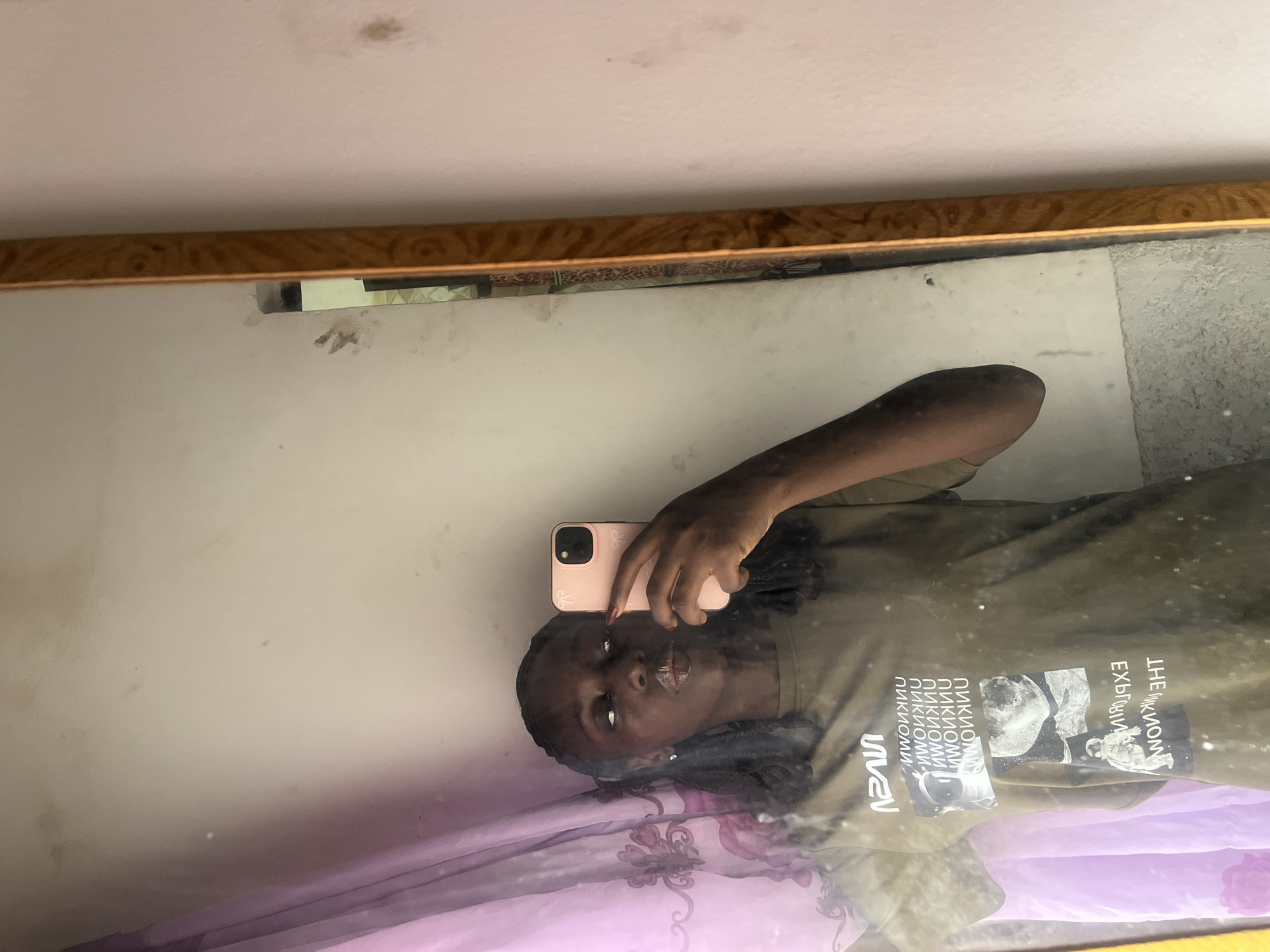 A person is taking a mirror selfie. The person has dark skin, dark hair, and is wearing an olive green t-shirt with a graphic design on the front. They are holding a pink iPhone to take the photo. The background includes a white wall, a wooden-framed mirror, and a pink and purple patterned blanket. The location is not discernible as Abuja, Nigeria, based on the visual information.