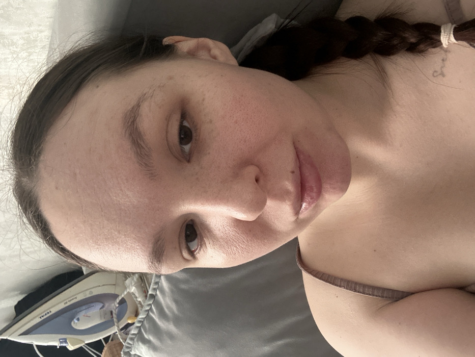 The image presents a close-up, eye-level indoor portrait of an adult female in Chaikovsky, Russia. The subject has light skin, dark brown hair pulled back with a visible braid secured by a light-colored elastic on her right side. Her brown eyes are directed towards the viewer, and her lips are slightly parted. A small mole is present superior to her upper lip. A faint, light-colored tattoo resembling text is discernible on her left clavicle area. She is attired in a light brown or beige thin-strapped garment. In the upper left background, a light blue or grey pillow is visible. Beyond the pillow, a white and blue Tefal electric iron is positioned upright on a light-colored surface, with a small yellow object partially visible nearby. The background wall is light-colored and appears textured.
