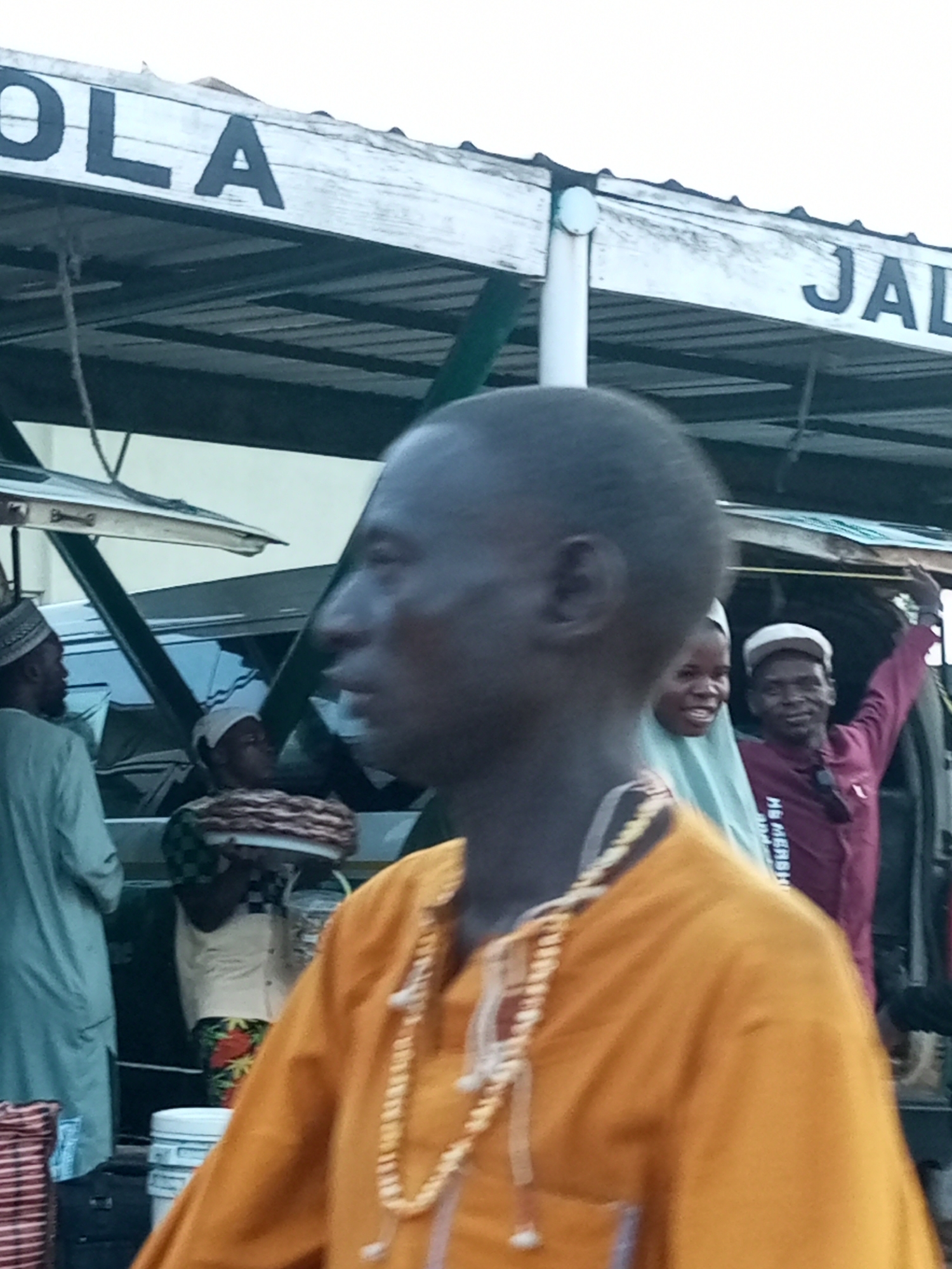 In Gombe, Nigeria, a man wearing an orange garment and a beaded necklace is pictured in profile facing right. Behind him, a woman in a light blue headscarf smiles. Further back, a man in a maroon jacket and a white baseball cap is visible. In the background, a sign reads "OLA" on the left and "JAL" on the right. The scene appears to be outdoors, possibly at a market or transport hub, with a corrugated metal roof overhead.