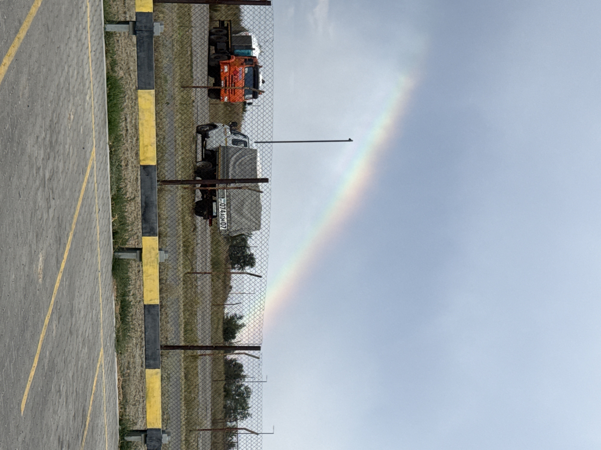 A rainbow arches across a pale blue, cloudy sky over a rural landscape. In the foreground, a chain-link fence separates a paved parking lot, marked with yellow lines, from a grassy area beyond. Two trucks are visible behind the fence: an orange truck on the left with a white tank on its bed, and a white truck on the right with a grey tarp covering its cargo area. The white truck has a license plate that appears to read "KZ 074 AGM 07". A tall, thin pole stands between the two trucks, and in the background, trees and more fencing can be seen. The scene is set in Приурал ауылдық округі, Kazakhstan.