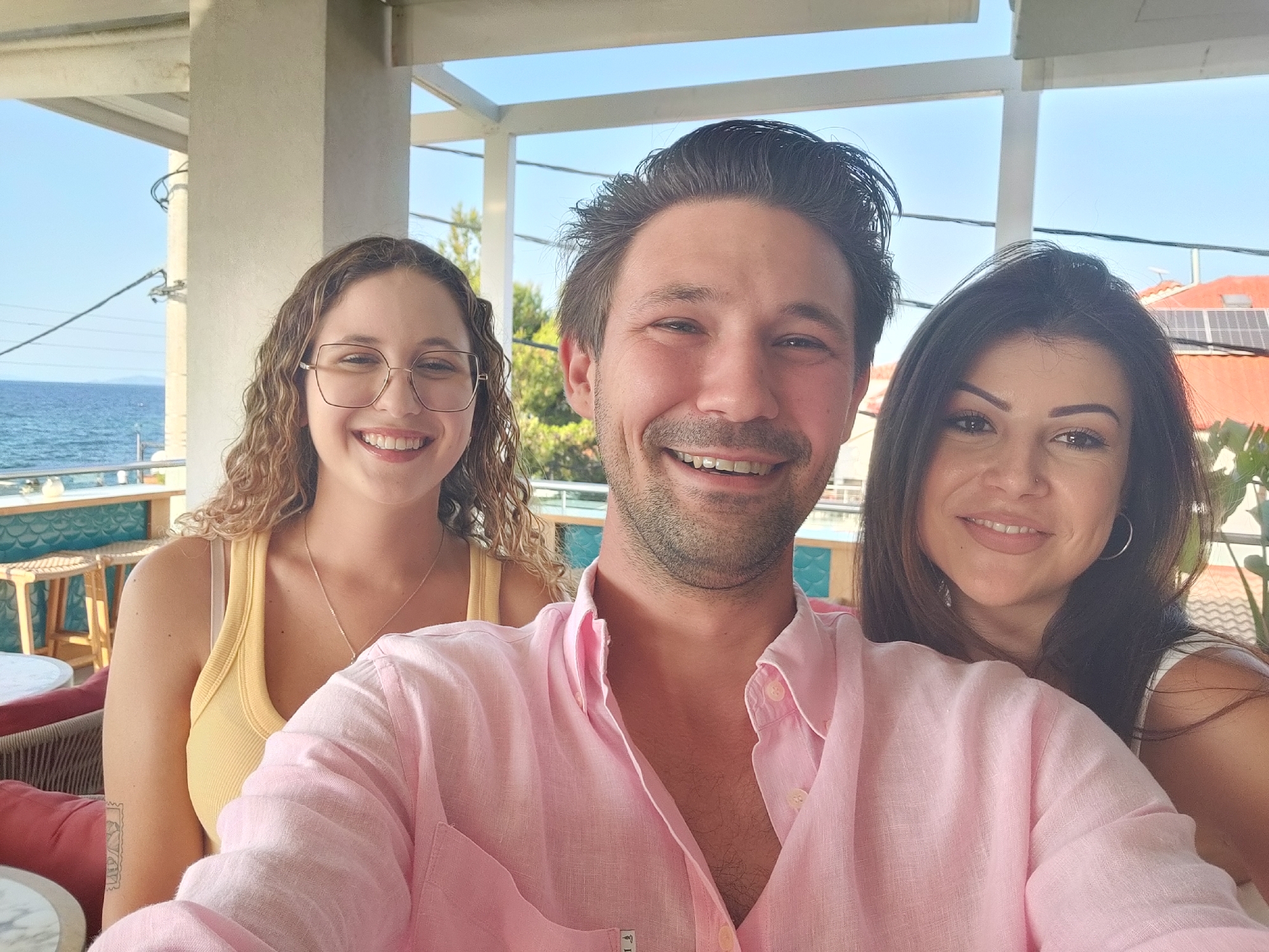 Three individuals, one male and two females, are captured in a selfie outdoors, likely on a balcony or terrace overlooking the sea. The man, positioned in the center, wears a light pink button-down shirt, unbuttoned at the collar, revealing chest hair. The woman to his left has curly blonde hair, wears a yellow tank top, and is seen with glasses and a necklace. The woman on his right has dark hair and wears a white top. All three are smiling at the camera. In the background, a bright blue sky and the azure sea are visible, with buildings and foliage suggesting a coastal urban or semi-urban setting. The scene appears to be in Sithonia Municipal Unit, Greece, given the coastal environment and architecture.
