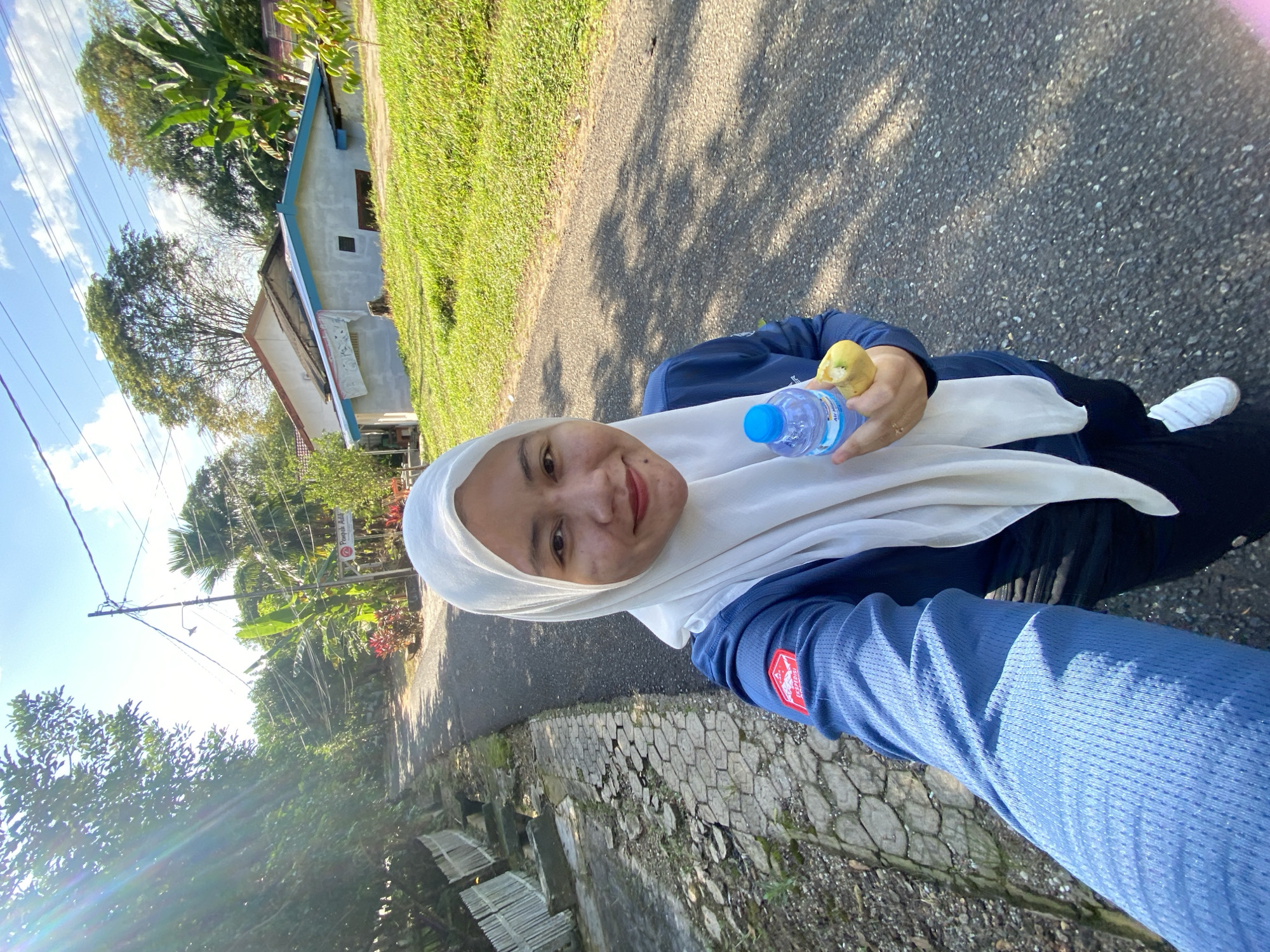A woman wearing a white hijab and a blue long-sleeved shirt is taking a selfie outdoors. She is holding a bottle of water and a partially eaten banana. The woman is walking on a paved road in a rural area. There are trees and houses in the background. The sky is blue with some clouds. The image was taken in Banjarbaru, Indonesia.