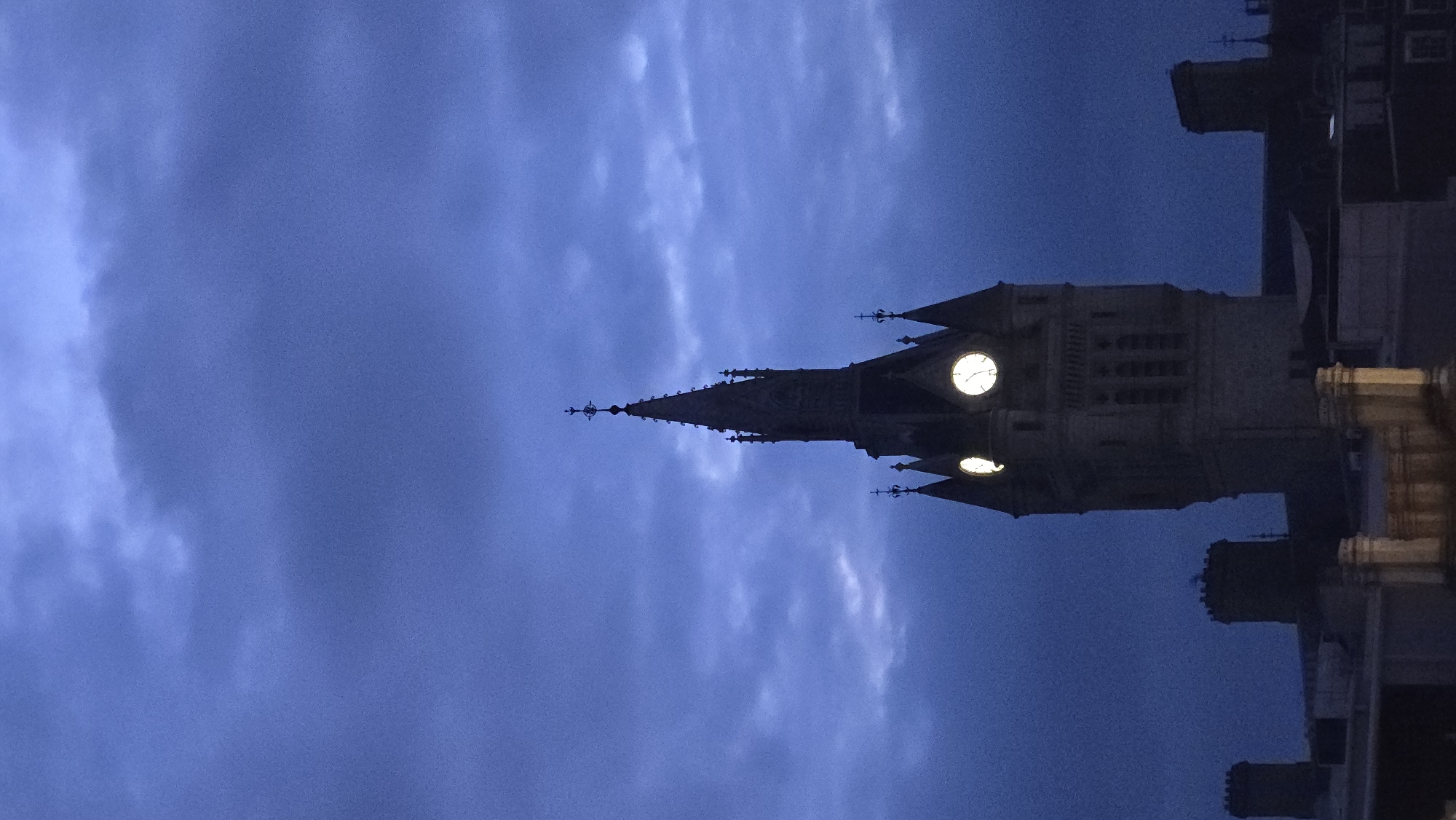 A tall, ornate clock tower dominates the center of the frame against a dark, cloudy sky. The tower, constructed of stone, features multiple levels, Gothic-style architectural details, and a pointed spire topped with a cross. Two illuminated clock faces are visible on the tower, appearing to be the primary source of light on its surface. Flanking the main tower are several smaller, less detailed structures, some of which resemble chimneys or turrets, and what appear to be rooftops of other buildings. The overall scene is dimly lit, suggesting it was taken at dusk or dawn, with the moody clouds contributing to a dramatic atmosphere. This is the Marischal College clock tower in Aberdeen City, United Kingdom.