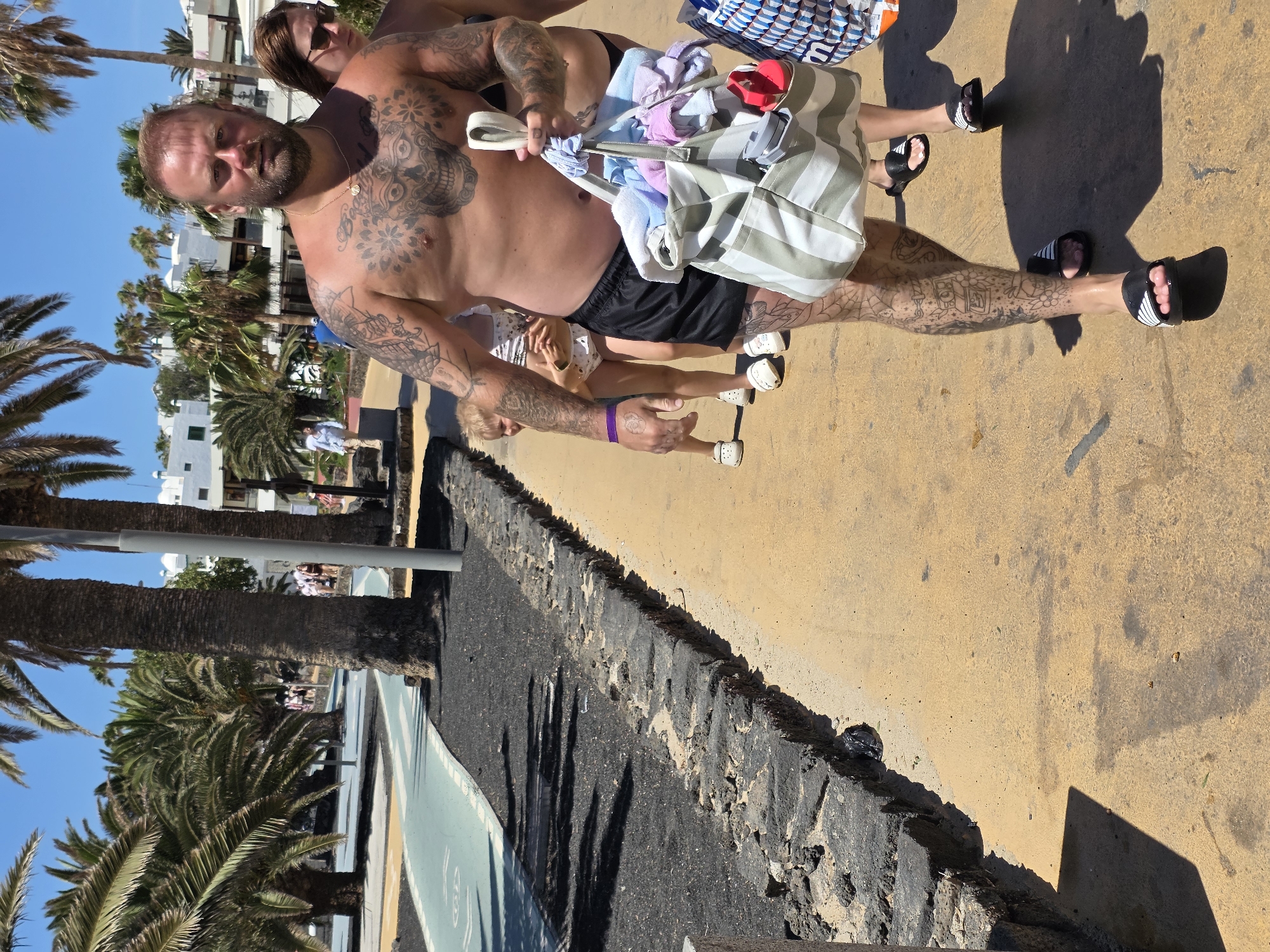 A man with full body tattoos and a beard walks towards the camera on a yellow paved pathway lined with dark volcanic rock. He is shirtless, wearing black shorts, and black and white Adidas slides. He carries a large striped tote bag filled with colorful towels and beach items. To his left, a young girl in a swimsuit and white Crocs is partially visible, followed by another woman in a bikini and sunglasses. Behind them, other people and palm trees suggest a tropical or coastal resort location. The background shows white buildings and more palm trees under a clear blue sky. The pathway appears to be a pedestrian walkway alongside a bike lane. The scene is lit by bright sunlight, casting distinct shadows. The location is identified as Teguise, Spain, likely a tourist area with volcanic landscape elements.