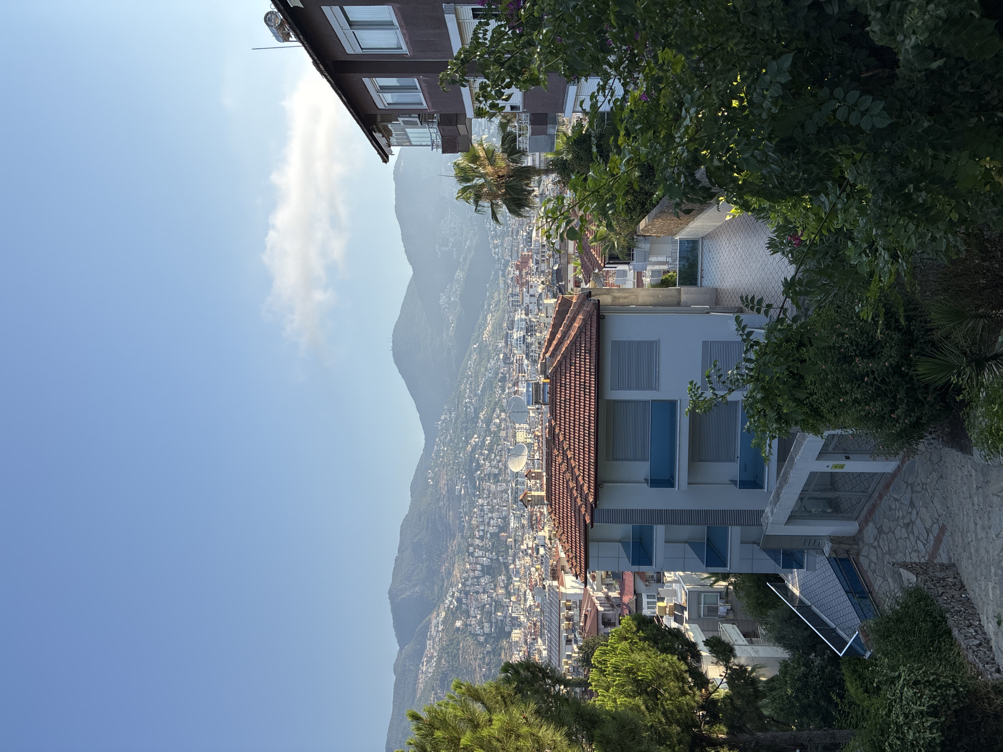 This image captures a daytime, elevated view of Alanya, Turkey, with a focus on residential buildings and a distant, mountainous cityscape. In the foreground, lush green trees and bushes frame the scene, with a stone pathway and staircase leading down to a courtyard area with a modern building. This building features white walls, blue-tinted glass balconies, and a tiled roof. To the right, a darker, taller building with multiple windows and balconies is partially visible.
Beyond the immediate structures, the middle ground reveals a dense urban landscape characterized by numerous buildings with red-tiled roofs, interspersed with other architectural styles. Solar panels are visible on some rooftops, indicating a likely modern residential area. Two prominent satellite dishes are mounted on a building in the mid-ground, suggesting television reception.
The background is dominated by the majestic Taurus Mountains, their slopes covered in a light haze and dotted with buildings that ascend the terrain. The clear blue sky, with a few wispy clouds, suggests fair weather. The overall impression is of a vibrant coastal city nestled against a dramatic natural backdrop.