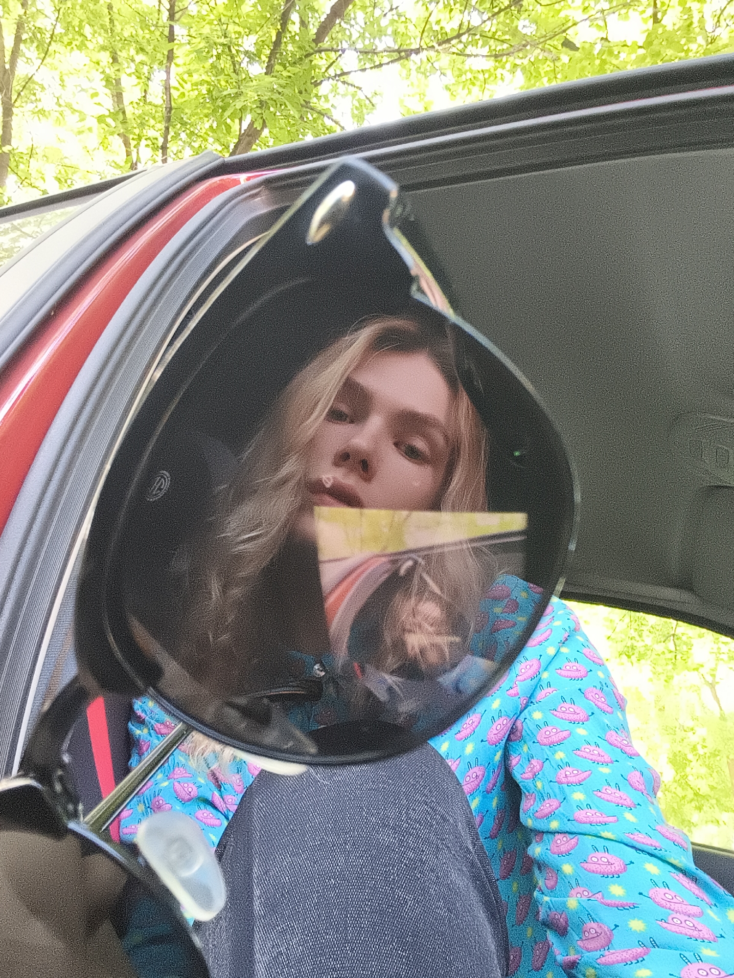 The image shows a person with long blonde hair sitting in a red car. The person is wearing a blue shirt with pink monster patterns and dark jeans. They are holding up a pair of black sunglasses, which reflects their face and parts of their clothing. The background includes green foliage, suggesting the car is outdoors, potentially near trees. The image lacks specific details to pinpoint the location within Izhevsk, Russia.