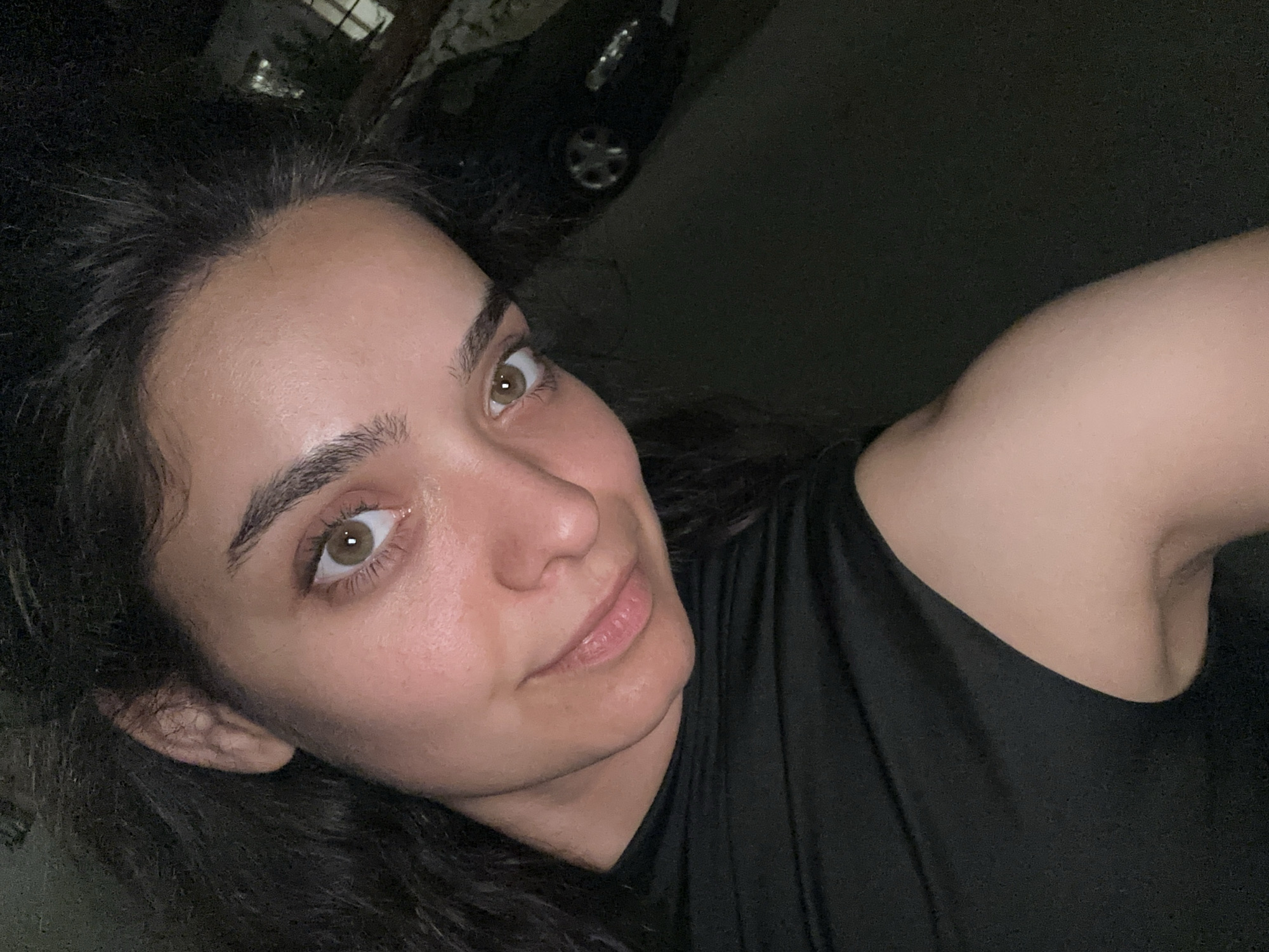 In a selfie taken at night, a woman with dark, curly hair and light brown eyes looks towards the camera. She is wearing a black top. Her expression is relaxed, with a slight smile. The background is dark, but a black car is visible to the right. The setting appears to be a residential area in Malevo, Bulgaria, suggested by the presence of a car and what looks like a building with lights in the distance.