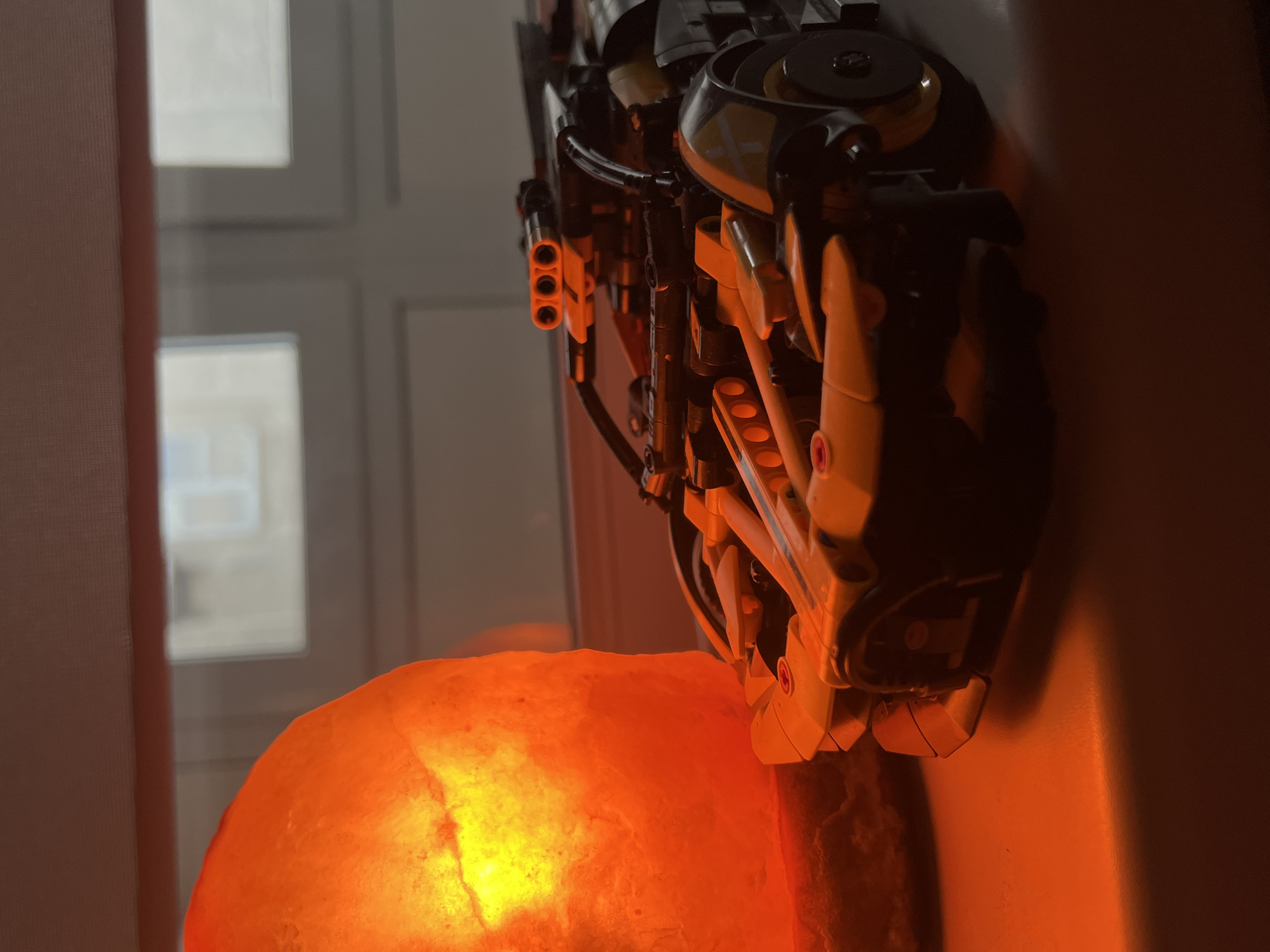 The image captures an indoor still life scene. In the foreground, a brightly glowing orange Himalayan salt lamp emits a warm light, illuminating its immediate surroundings. Above the salt lamp, a complex, mechanical-looking object is prominent. This object appears to be a model or toy, possibly made of building blocks like Lego Technic, with components in black, dark grey, and yellow/tan. It features intricate details such as tubing, gears, and a section with three small, cylindrical barrels or nozzles pointing leftward.
Behind these main subjects, a window is visible, offering a blurred view of an exterior environment, possibly showing other buildings or architectural elements. A light-colored curtain or blind partially frames the left side of the window. A plain wall forms the background on the right. The lighting suggests that the room is otherwise dim, with the salt lamp providing a significant source of illumination. There is no discernible activity taking place, nor is any text visible in the frame.