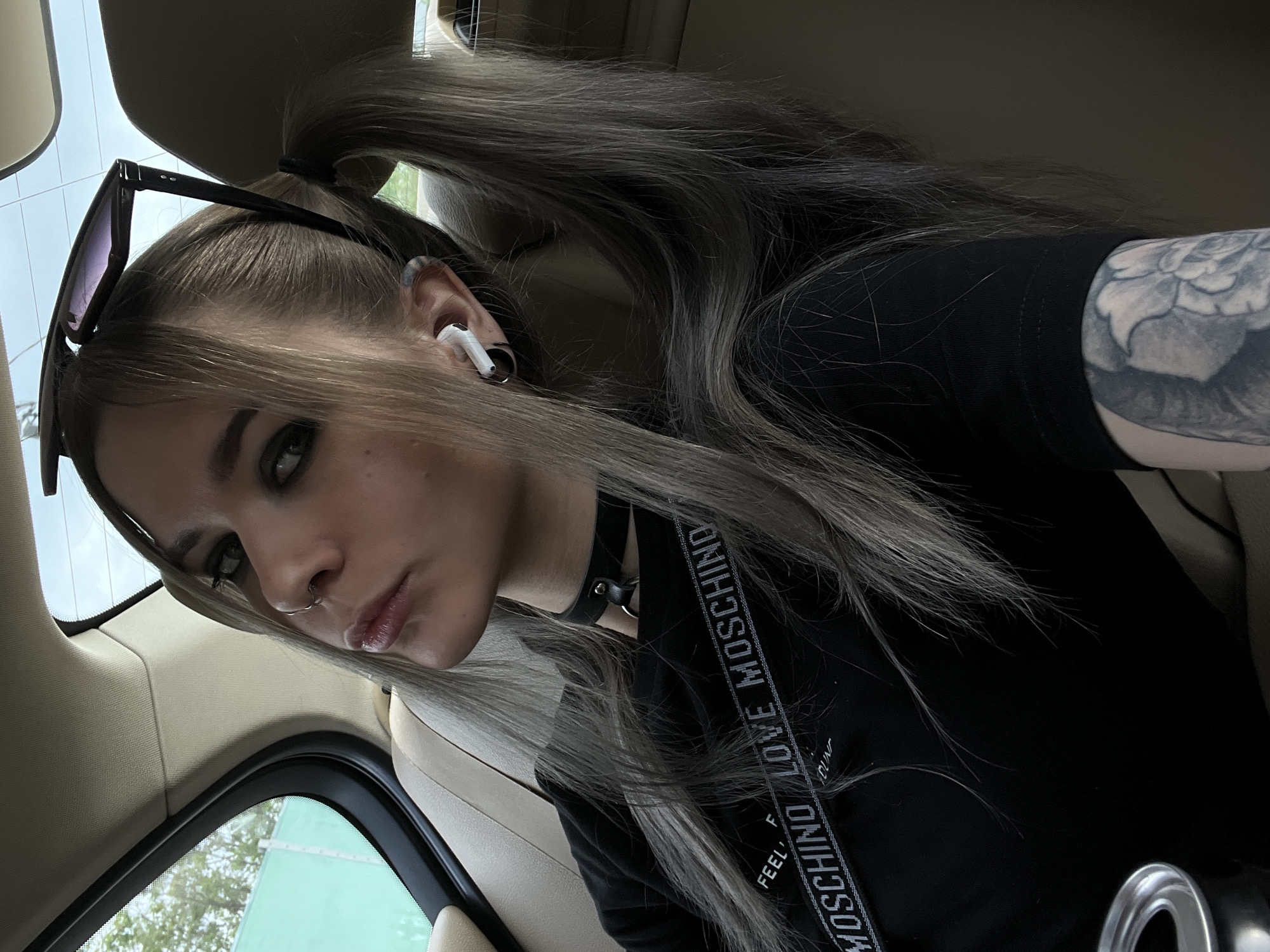 A young woman is photographed from a low angle inside a vehicle. She has light brown and grey hair partially tied up in a high ponytail with some strands framing her face. She wears a black choker and a black shirt with a "MOSCHINO LOVE MOSCHINO" strap. Her left arm is visible and has a tattoo. Sunglasses are perched atop her head. An earbud is in her left ear, and she has a nose ring. The interior of the vehicle is a light beige color. Through the vehicle's window, a glimpse of green foliage is visible. The location is not discernible as Khimki, Russia based on the visual information provided.