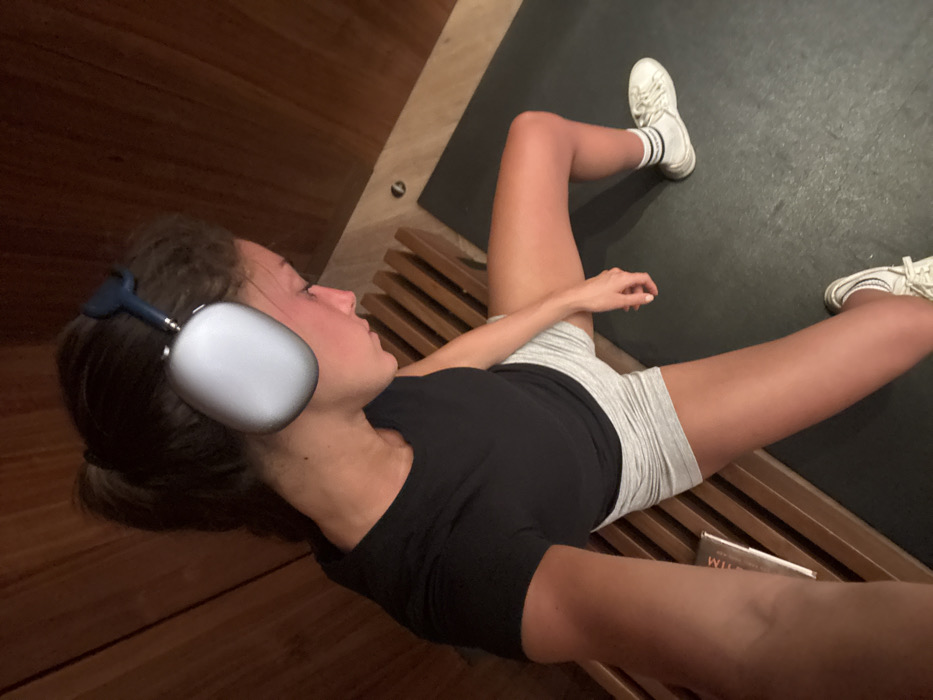 A woman lies on a wooden bench with her legs spread, wearing white sneakers with black stripes. She has on a black t-shirt and gray shorts, and white headphones with blue accents rest on her head. A black mat lies next to the bench, and a book sits on the bench between her legs. One arm is visible in the foreground. The photo is taken from a high angle.