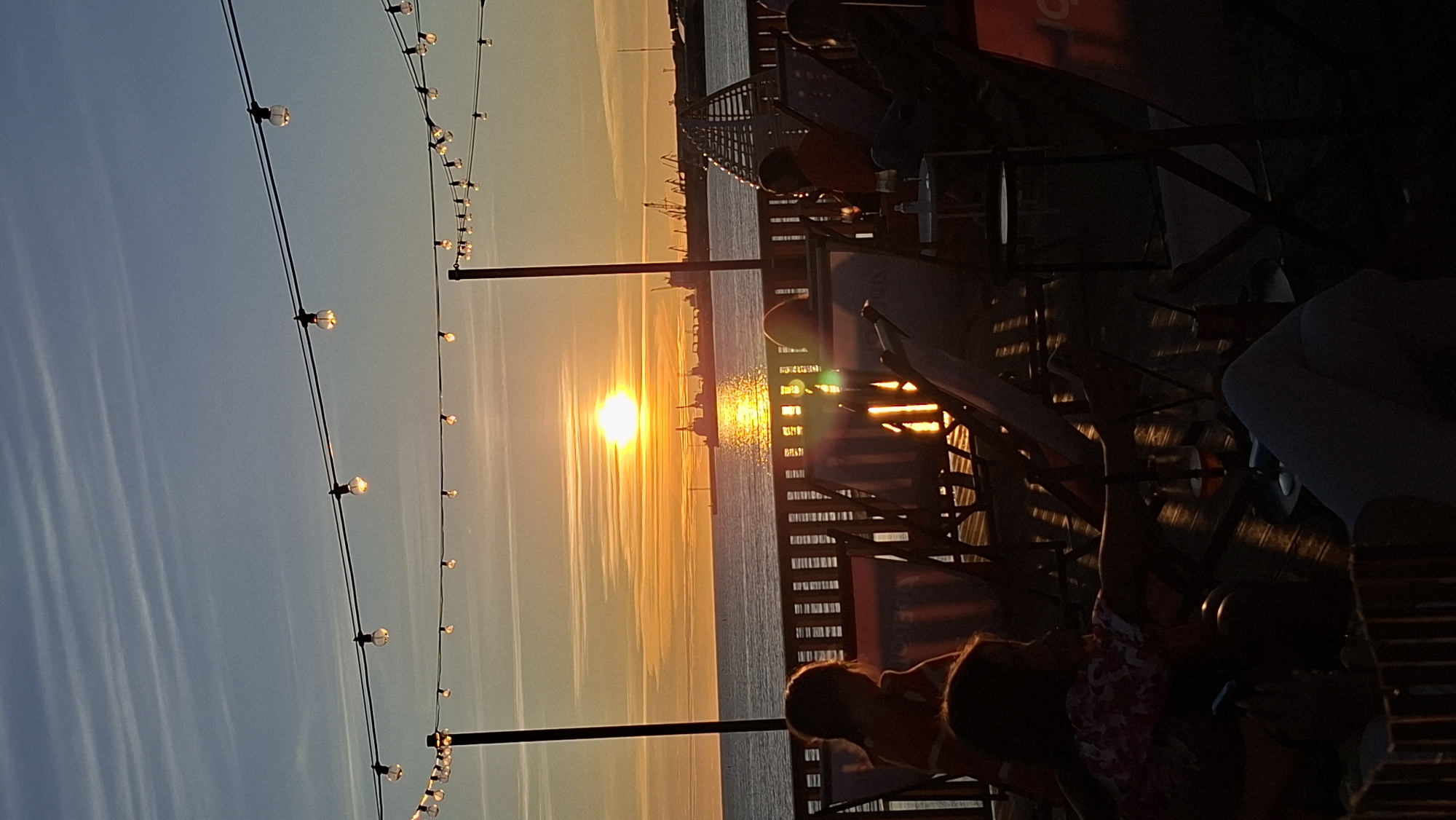 An evening scene at a waterfront venue in Tallinn, Estonia, features a sunset over the Baltic Sea. The sun, a brilliant orange orb, is setting on the horizon, casting a shimmering golden path across the water. Silhouetted against the warm glow are several cargo ships and industrial structures in the distance. In the foreground, people are seated in chairs on a wooden deck or pier, some facing the sunset. String lights with exposed bulbs are suspended overhead, adding to the ambient atmosphere. The overall impression is one of a relaxed evening gathering by the sea.