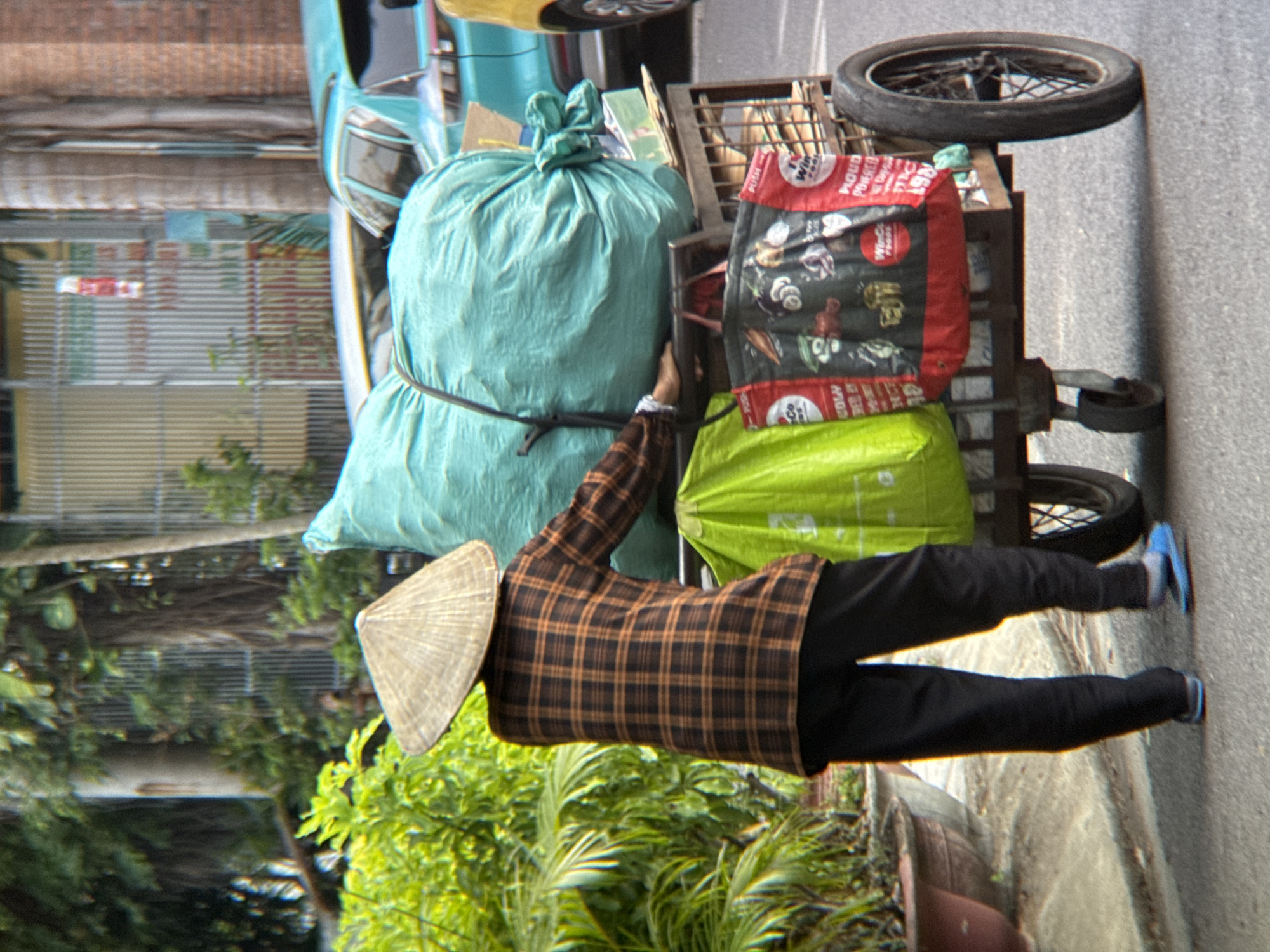 A person wearing a conical hat and a plaid shirt is pushing a cart laden with large bags and other items. The cart has at least three wheels, two large ones on the back and one smaller one in the front, and appears to be made of metal. One of the large bags on the cart is a bright green, and another is a large, light blue bag. There is also a red and black patterned bag with white lettering and images of food items. The person's lower half is visible, wearing black pants and blue flip-flops. Behind the person and cart, a teal-colored car is parked. The scene appears to be on a street, with a sidewalk and some greenery visible on the left. The overall impression is that of a street vendor or someone transporting goods. This scene is located in Nha Trang, Vietnam, indicated by the presence of conical hats, typical street vending carts, and the general urban environment consistent with Southeast Asian cities.