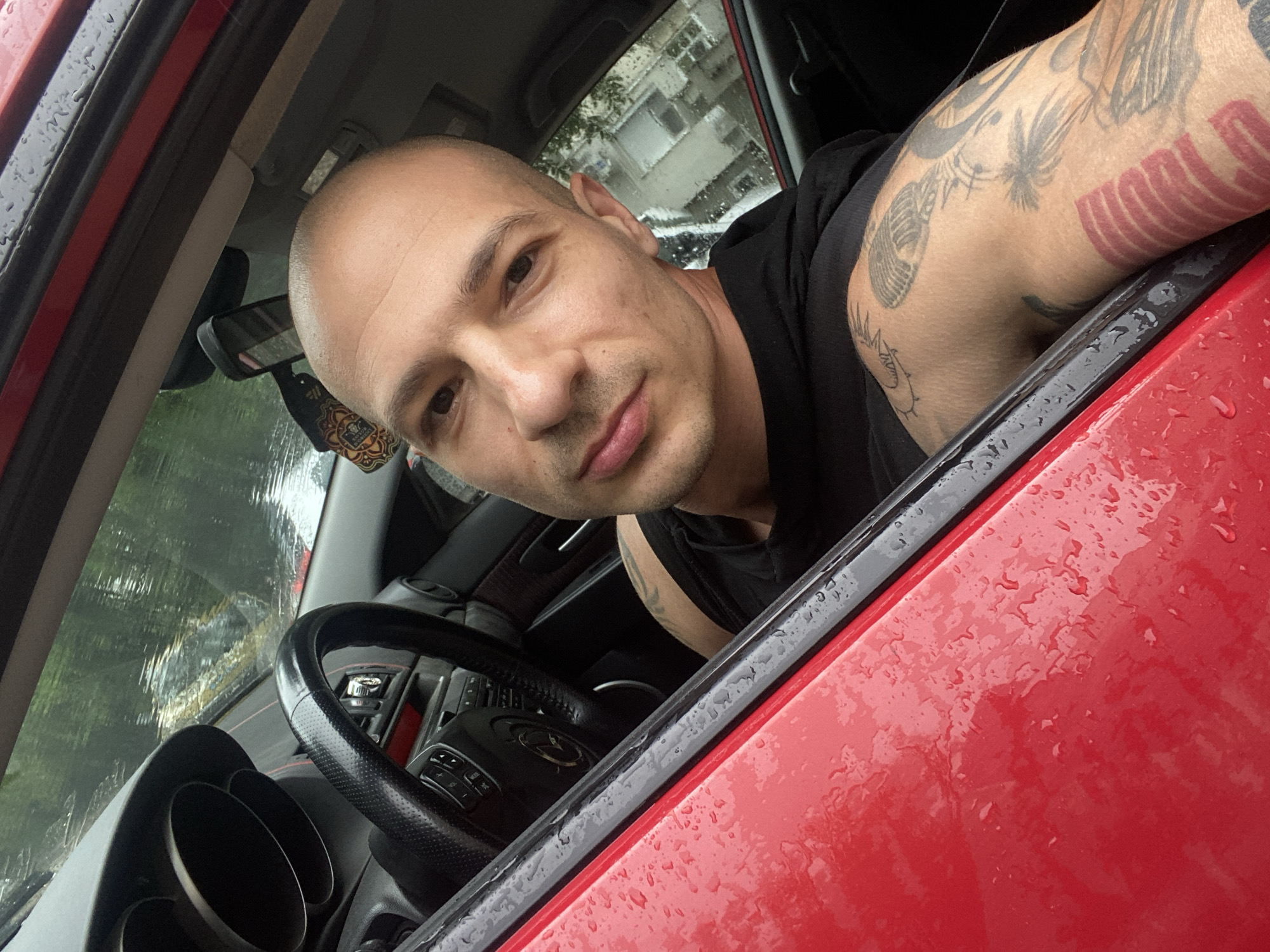 A close-up, slightly angled shot features a man seated in the driver's seat of a red car, likely in Pleven, Bulgaria. The man, appearing to be in his 30s or 40s, has a bald head and short facial stubble, looking directly at the camera with a neutral expression. His right arm, which has various tattoos including a microphone, foliage, and red block letters, rests on the car's open window frame. He is wearing a black sleeveless top.
The setting is the interior of the car, with parts of the black dashboard, steering wheel, and a rearview mirror visible. An air freshener with a detailed, mandala-like design hangs from the rearview mirror.
A significant detail is the weather: numerous raindrops are clearly visible on the red exterior paint of the car door and the window, indicating it is raining or has recently rained. Through the wet window, blurred reflections of buildings and trees can be seen, suggesting an urban or suburban environment. The overall lighting indicates it is daytime. No clear, readable text is visible on any objects or surfaces within the frame.