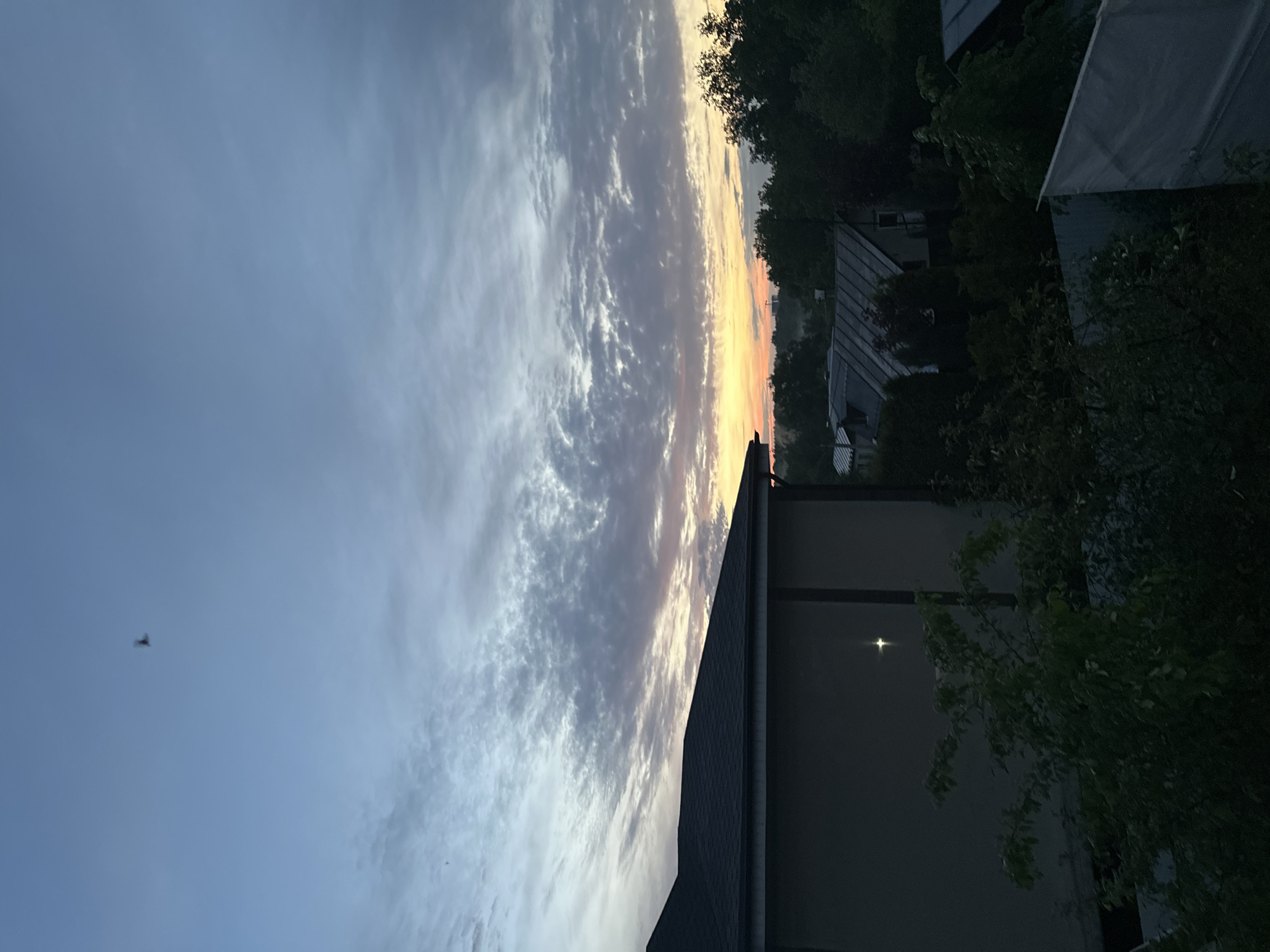 The image shows a dusk sky with a band of orange light at the horizon. Above the horizon is a large area of streaky gray and white clouds, and higher still is a clear, light-blue sky. In the lower right of the frame are the eaves and roof of a building, with the side of the building and tree branches extending out from behind the building. Other buildings and more trees are visible at the horizon. In the bottom left of the frame is a small bird in flight. I have no evidence to suggest this image was taken in Nizhny Novgorod.