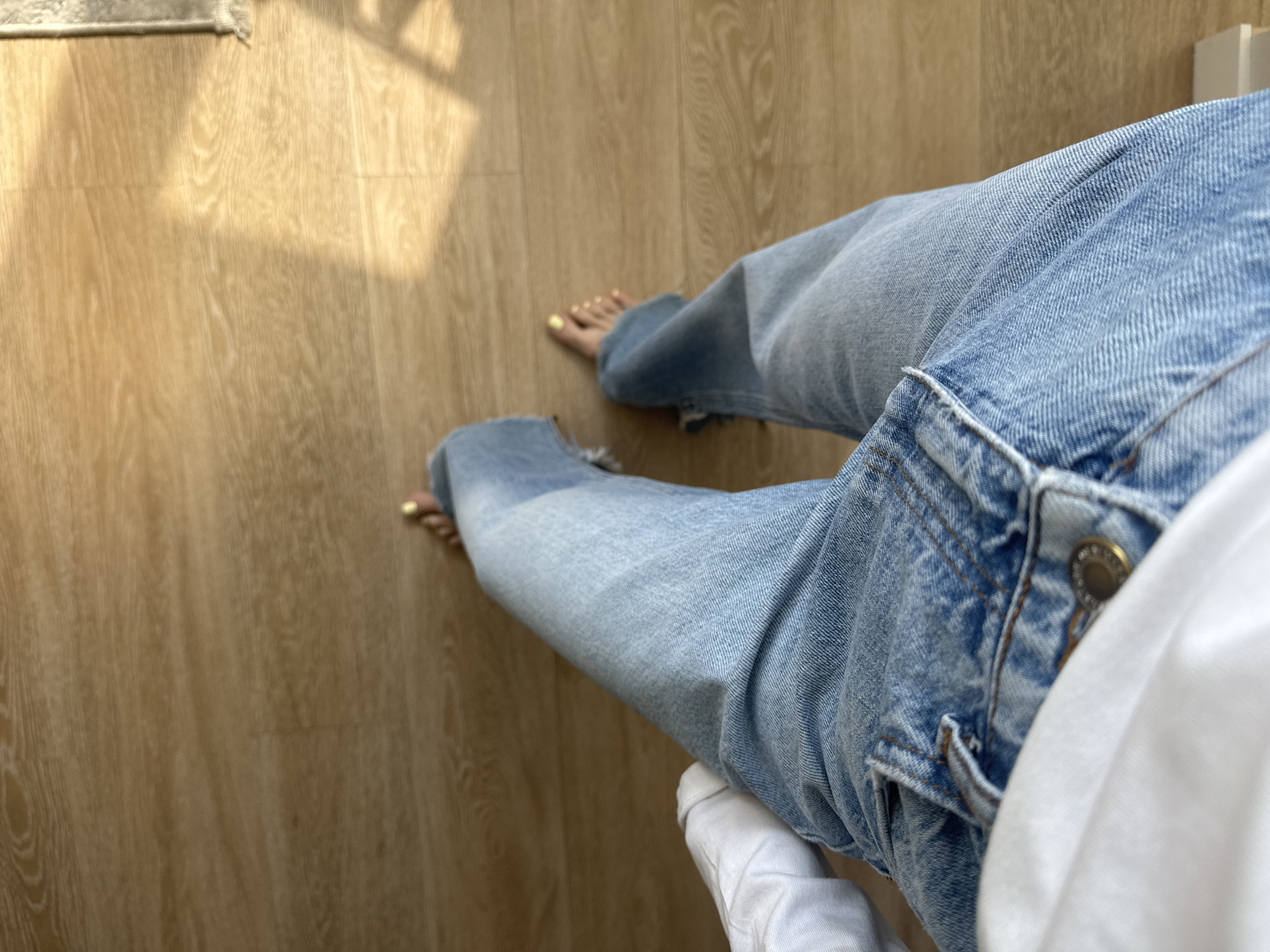 The image shows a view looking down at a person wearing light blue denim jeans sitting on a white cloth-covered surface. The person is barefoot, with visible toenails painted yellow. The background is a light brown, wood-grain patterned surface, possibly a floor. Sunlight is casting shadows on the floor. The location is not specified in the image and there is no indication of it being in Moscow, Russia.