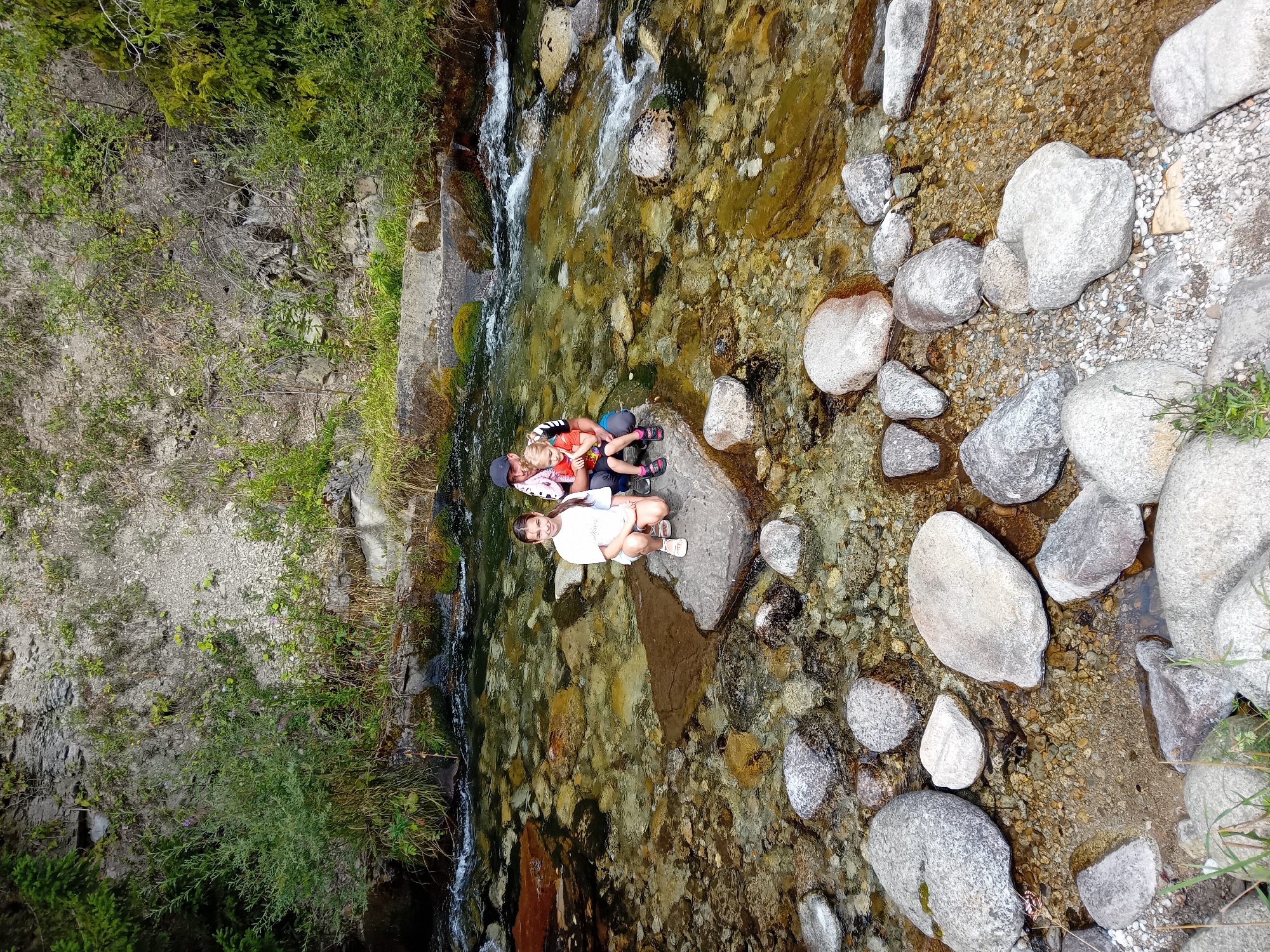 In Zuberec, Slovakia, three individuals, an adult and two children, are positioned on rocks within a shallow, clear stream. The adult, wearing a dark baseball cap and a pink shirt, is seated, holding a younger child on their lap. The younger child is wearing an orange shirt. To the left, a taller girl in a white t-shirt and shorts is crouched on a separate rock. The stream bed is visible, filled with numerous smooth, multicolored rocks and pebbles, and the water flows gently over them. The background consists of a steep, rocky embankment with sparse vegetation, including bushes and trees. The overall environment suggests a natural, outdoor setting, likely a mountain stream or river.