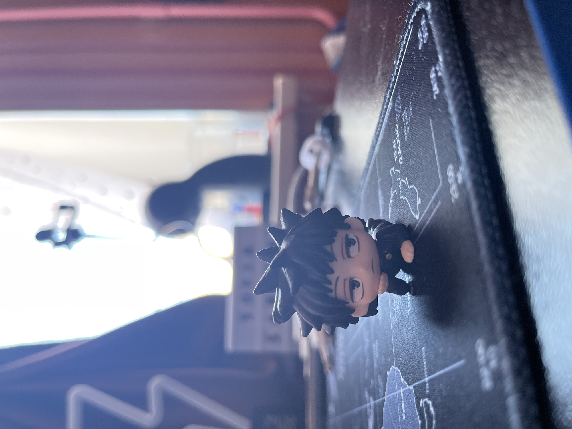 A small figurine of an anime character is lying on a textured black surface, possibly a mousepad with a world map design. The figurine has spiky black hair, large eyes, and is wearing a dark uniform with a gold button. Its head is tilted to the side, and its mouth is slightly open. The background is out of focus and appears to be a room with bright light coming from a window, casting a blue and white glow on a dark blue surface. The overall lighting suggests it is daytime. The figurine is the primary subject, placed centrally on the black surface. The scene appears to be a personal desk or workspace. No text is clearly legible.