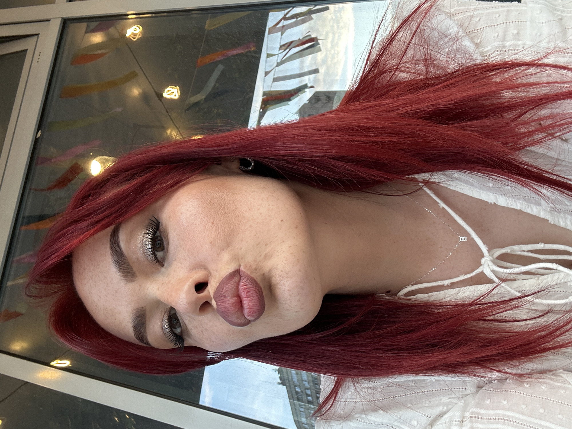 A close-up selfie shows a woman with bright red hair and freckles, pouting her lips. She is wearing a white, textured blouse with a tie detail at the neck and a delicate silver necklace with a small charm. Her eyes are made up with eyeshadow, eyeliner, and mascara, and she wears a small hoop earring in her left ear. She is positioned in front of a window with a reflection of a city skyline, possibly in Tolyatti, Russia, given the context of the request. Decorative lights and colorful bunting are visible through the window, suggesting an outdoor or semi-outdoor setting like a cafe or restaurant.