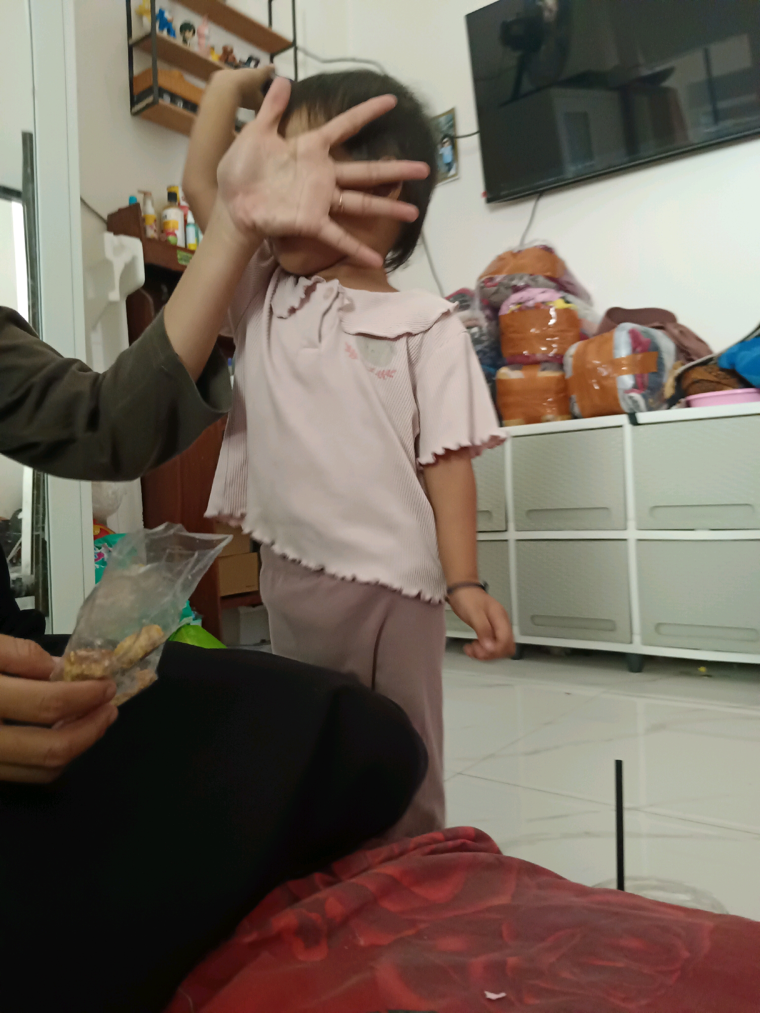 The image depicts an indoor scene, likely a living room or bedroom, within a residence in Sragen, Indonesia. A young child, facing away from the camera and partially obscured by their hand, stands in the center of the frame. The child is wearing a pale pink t-shirt with ruffled sleeves and light pink pants. Their left arm is raised, and their hand is held up in front of their face, obscuring it. To the left of the child, another individual, whose arm is visible, is holding a clear plastic bag containing small, light-colored items, possibly snacks or treats. The background reveals a wall with a television mounted on it, a shelf with various items, and a white cabinet with multiple drawers. The floor is tiled, and a red blanket or piece of fabric is visible in the foreground. The overall impression is a casual, domestic moment, potentially interrupted by the act of taking a photograph.
