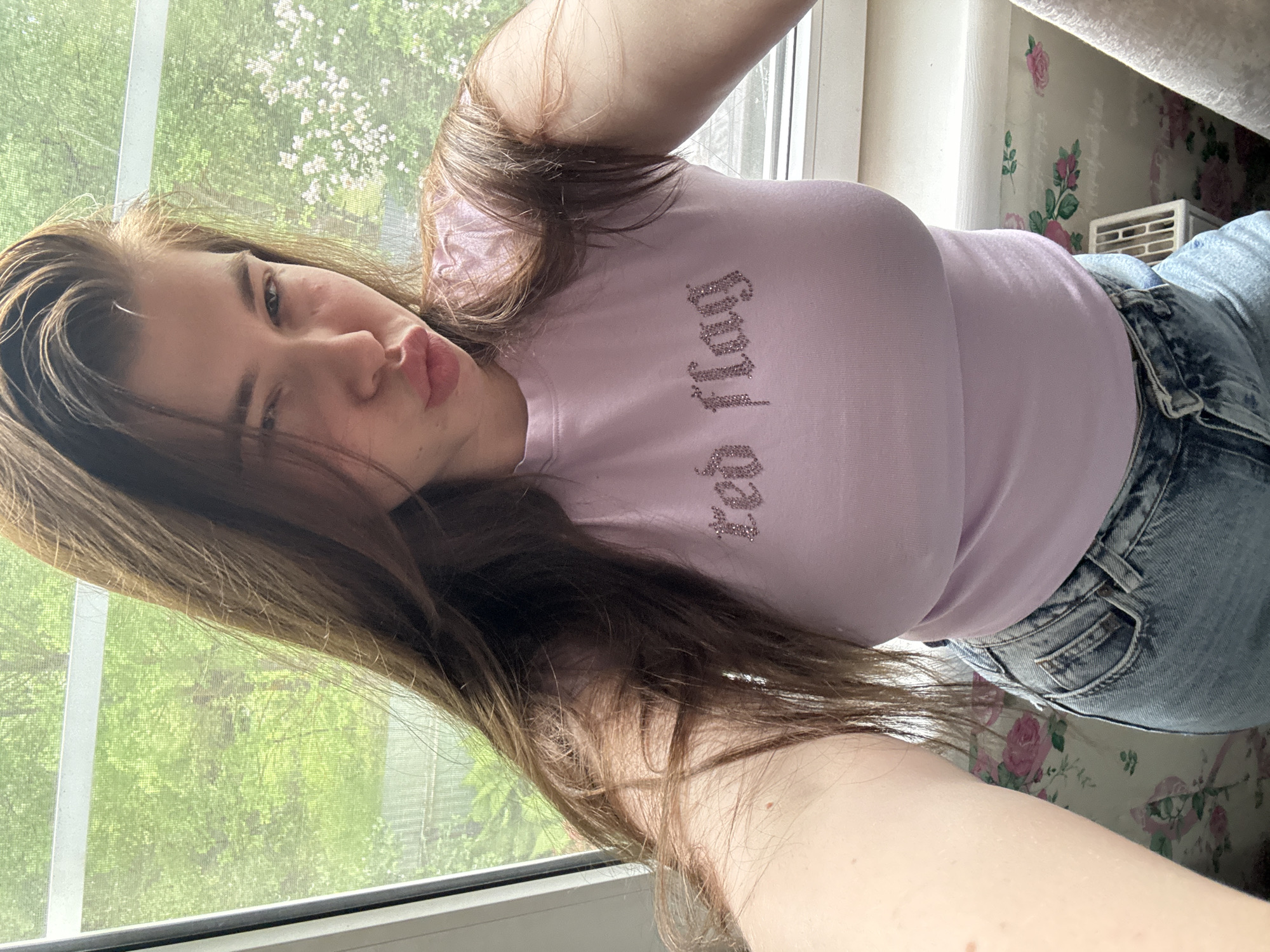 A young woman with brown hair and fair skin is taking a selfie. She is wearing a light purple t-shirt with the word "Juicy" written on it, and light blue denim jeans. She is winking and puckering her lips. Behind her is a window with a view of trees and greenery. There is also a wall with a floral pattern behind her. The location does not appear to be in the city of Meshchery, Russia based on visual indicators.