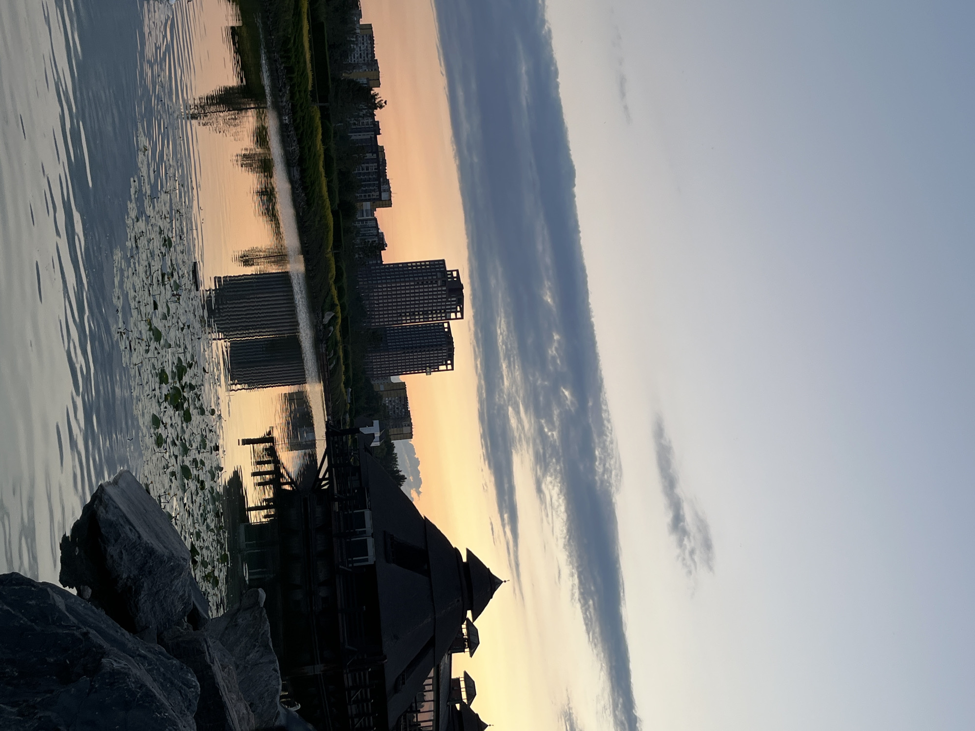 The image depicts a waterfront scene at dusk, likely in Dnipro, Ukraine. In the foreground, large rocks are piled along the shore, hinting at a breakwater or natural embankment. A dark, multi-tiered structure with a peaked roof, possibly a pavilion or boathouse, occupies the lower right portion of the frame, its silhouette stark against the fading light. Beyond this structure, a body of water reflects the sky and the distant urban landscape.
The water is dotted with lily pads, suggesting a lake, river, or canal. Across the water, a line of buildings, including several modern high-rise apartments, is visible. The sky exhibits the warm hues of sunset, transitioning from soft oranges and yellows near the horizon to a pale blue overhead. Wispy clouds stretch across the sky, some appearing dark and dramatic, others thin and elongated. The overall mood is serene and tranquil, characteristic of a quiet evening. The reflections in the water are slightly distorted by ripples, adding a dynamic element to the stillness. There are no people visible in the image, and no text is present. The scene conveys a sense of urban life coexisting with nature, viewed during a peaceful time of day.