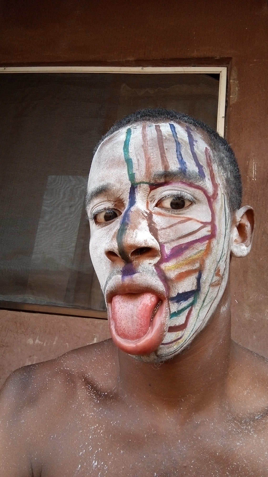A person with dark skin and short, dark hair is seen from the chest up. The person's face is painted white, with colorful lines and patterns on the right side. The person's tongue is extended out of their mouth. The background includes a window with a screen and a reddish-brown wall. The location is Onitsha, Nigeria.