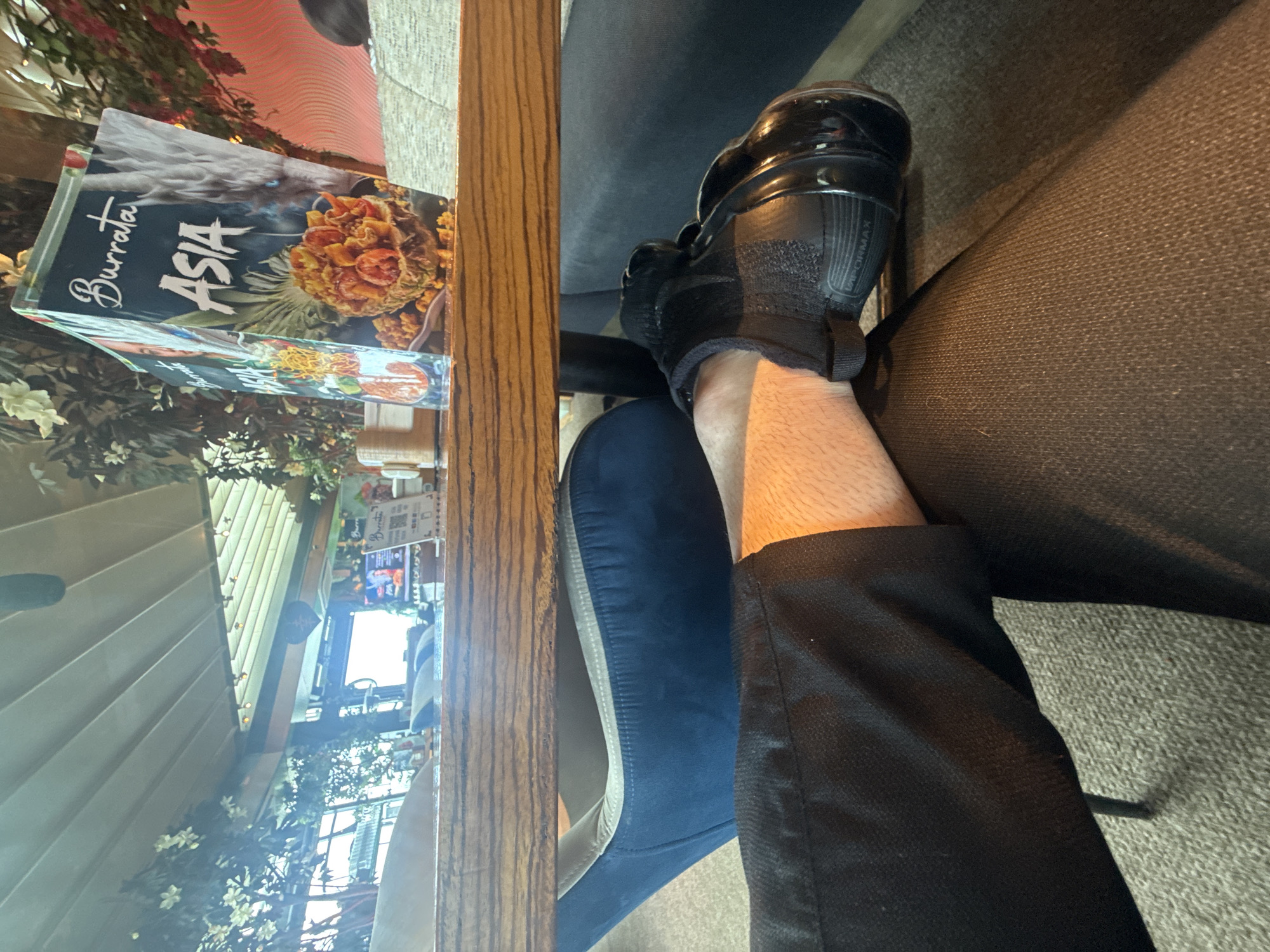 The image presents a low-angle interior view from a restaurant, specifically "Burrata Italiana & Asia" located in Paradise Center, Sofia, Bulgaria. In the foreground, a person's legs are depicted. The left leg, clad in dark trousers, reveals a section of bare, fair-skinned shin. The right leg, also in dark trousers, is crossed over the left, with the foot wearing a black Nike Vapormax sneaker, identifiable by text on the heel. The floor is covered with light-colored carpet. Above the legs, a wooden table surface with distinct grain is prominent. On the table, two menus for "Burrata ASIA" are displayed, featuring food imagery and a stylized dragon design. A small, light-colored table accessory, possibly a napkin holder, is also visible. Beneath the table, a blue upholstered chair is partially observed. The background reveals a well-lit interior space with large windows allowing natural light. The ceiling exhibits a white, slatted, possibly retractable structure. Artificial foliage, including white flowering branches and green vines, decorates the setting. Distant background elements include additional seating and a faint television screen displaying colorful graphics. No direct human interaction or faces are visible.