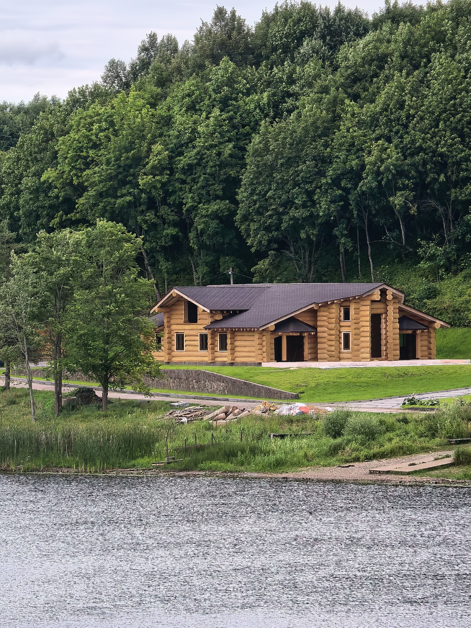 The image depicts a large, modern log cabin situated on a grassy slope overlooking a calm body of water. The cabin is constructed from substantial logs, featuring a dark, pitched roof, multiple windows, and open doorways. The surrounding environment is lush and green, with a dense forest of deciduous trees forming a backdrop to the cabin and a line of reeds growing along the water's edge. In the foreground, the rippling surface of the water reflects the overcast sky. Construction materials, including stacked lumber and what appear to be bags of sand or cement, are visible near the water's edge. The scene is one of a rural or semi-rural setting, likely a residential or recreational property. The location is identified as Staraya Ladoga, Russia. There are no people visible in the image.