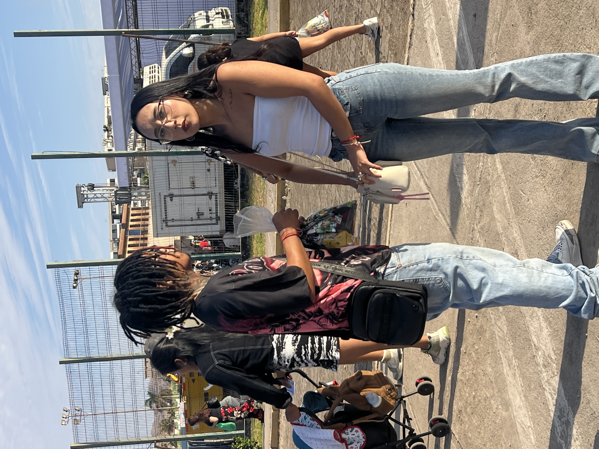 The image depicts a street scene in Arica, Chile, with a focus on two young individuals. The person in the foreground on the left has dreadlocks and is wearing a black t-shirt with a graphic print and light blue jeans, carrying a black crossbody bag. They are holding a clear plastic bag. To their right, a young woman with glasses and long dark hair is wearing a white strapless top and light blue flared jeans, holding a white purse. They appear to be interacting, possibly in conversation. In the background, a metal fence encloses an area where a white truck and other structures are visible, suggesting an event or a specific venue. A stroller is partially visible to the left of the dreadlocked individual. The ground is paved, and the sky is a clear blue with scattered clouds, indicating daylight. Other people and objects are present in the background, contributing to a sense of an outdoor gathering or public space.