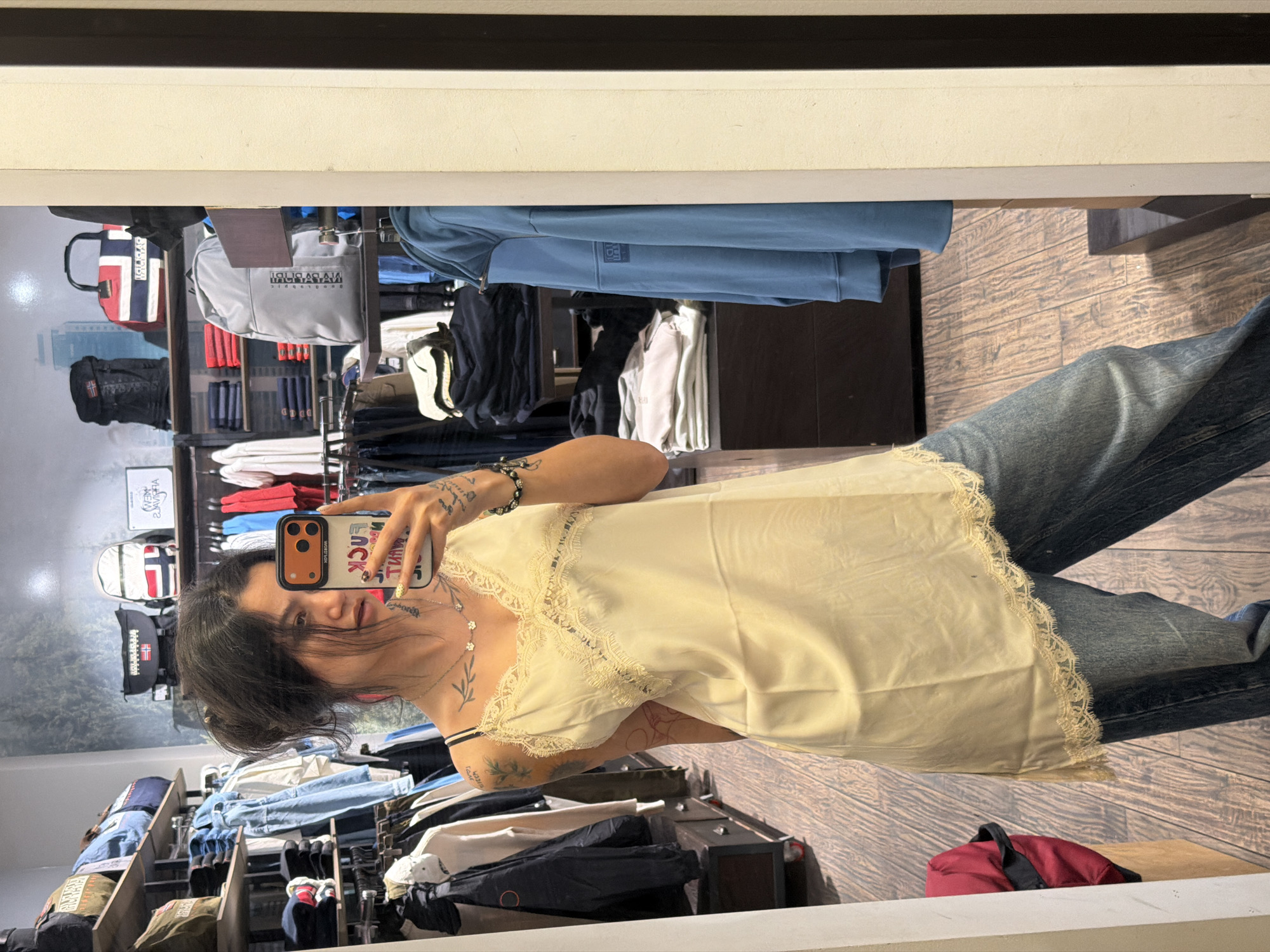 A woman is taking a picture of herself in a mirror in a clothing store. The mirror is reflecting a clothing display rack with various garments, including jackets, shirts, and sweaters. The woman is wearing a light yellow top with lace trim and blue jeans. She is holding a mobile phone with a protective case. The image's location is not explicitly shown to be Sofia, Bulgaria, and cannot be determined from the scene.