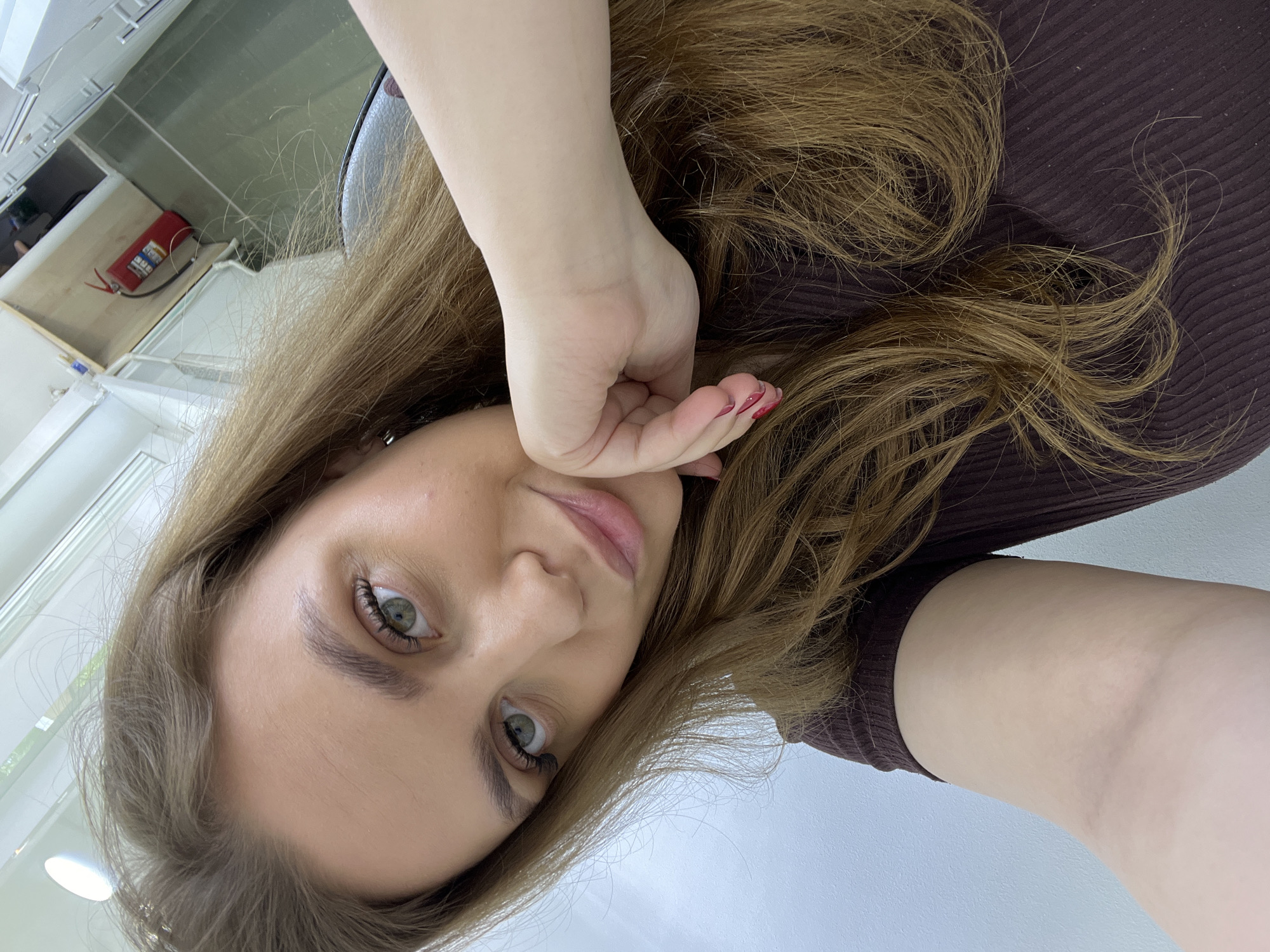 A woman with long brown hair is facing the camera. She is wearing a brown ribbed top. Her left hand is resting on her face. The background appears to be an interior space, possibly a salon, with a white wall and what looks like a fire extinguisher. The image is rotated 90 degrees. There is no indication this is in Chaikovsky, Russia.