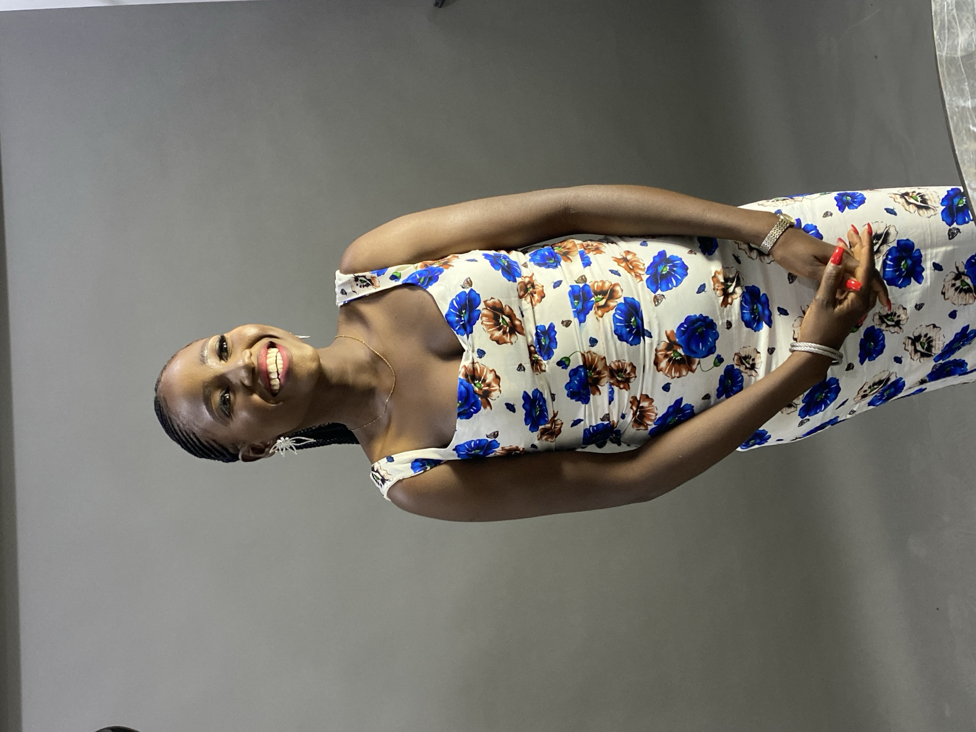 This portrait features a young woman with dark skin, captured smiling warmly against a plain, solid grey background. Her hair is styled in small, neat braids, visible along the sides of her head. She is looking directly at the viewer with a wide, joyful smile, revealing her white teeth and pink gums.
She is dressed in a sleeveless, knee-length dress with a white base, intricately patterned with large floral designs in vibrant blue, earthy brown, and subtle black or grey tones. Her accessories include a delicate gold-colored chain around her neck and distinctive spider-shaped earrings. On her left wrist, she wears a classic metallic watch, while her right wrist is adorned with a silver-colored bracelet. Her fingernails are painted a striking red, and her hands are clasped gently together in front of her midsection.
The background appears to be a simple studio backdrop or an interior wall, providing a clean and uncluttered setting. The lighting is soft and even, suggesting an indoor environment. The image conveys a sense of happiness and confidence from the subject. There is no visible text. The context is a posed photograph.