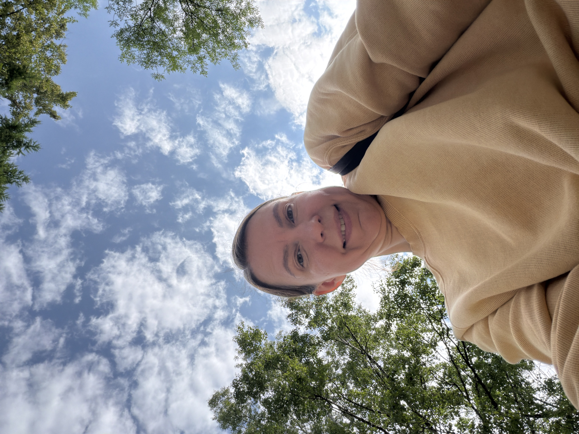 A low-angle, first-person perspective selfie shows a woman with her head tilted back, looking up towards the sky. She has blonde hair pulled back and is smiling. She is wearing a light brown sweatshirt and a black strap is visible over her shoulder. The background is a bright blue sky with scattered white clouds, and the tops of green trees are visible around the edges of the frame. The image was captured in Chaikovsky, Russia, likely in a park or natural area given the presence of trees.