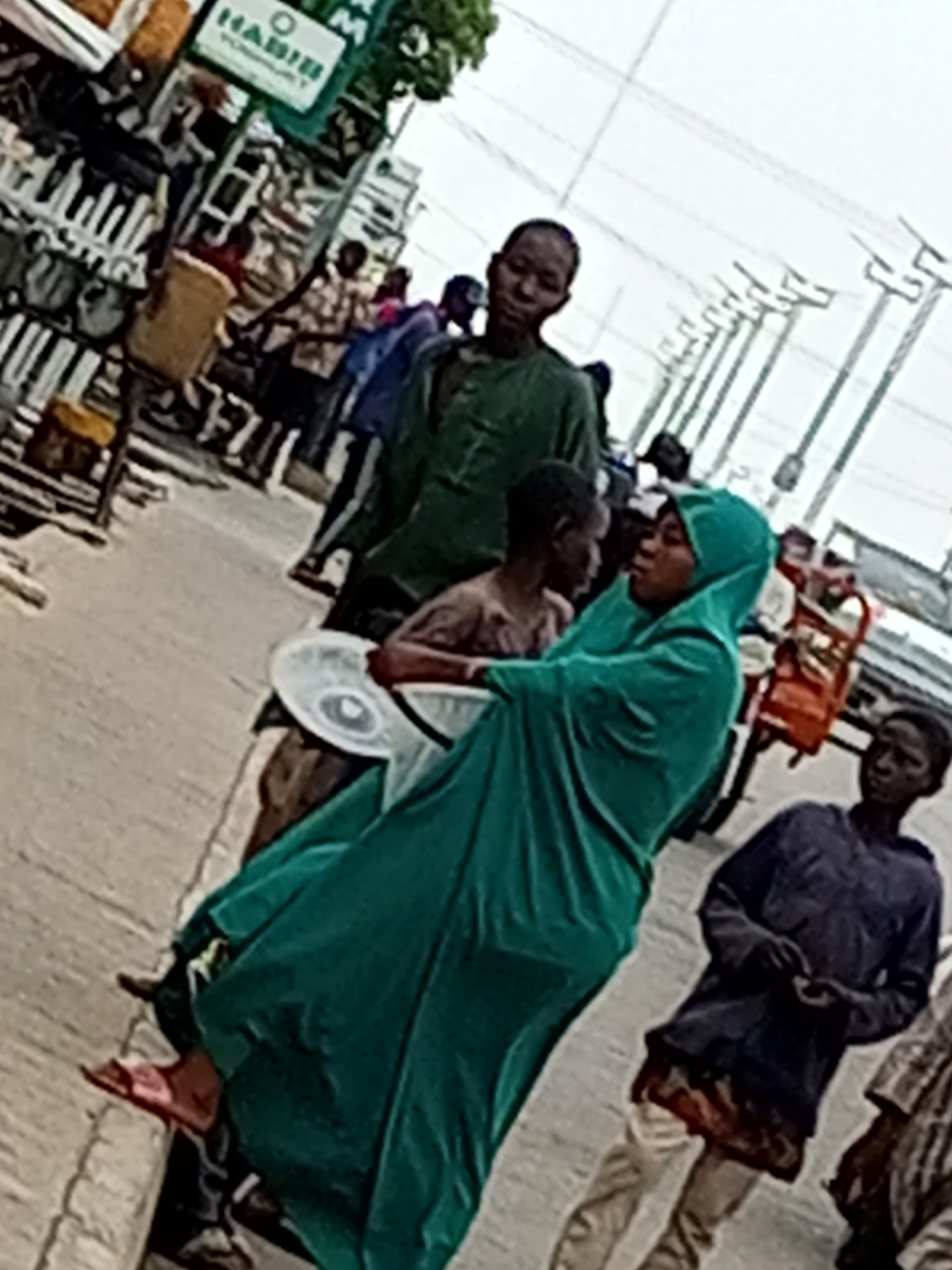 This is a low-resolution, slightly blurry, wide shot of a street scene in Tumfure, Nigeria. In the foreground, a person in a green hijab is walking, carrying a white circular object. To their left, a young boy with a bare torso walks beside them. Behind them, several other individuals, likely adults and children, are visible in the midground and background. Some are wearing casual clothing, while others appear to be in traditional attire. A green sign with "HABIB" written on it in white lettering is visible on a pole to the left of the frame, suggesting a business or street vendor. The environment appears to be an outdoor market or street with a paved ground. Overhead power lines are visible in the upper portion of the image. The overall impression is a candid snapshot of daily life on a street in Nigeria.