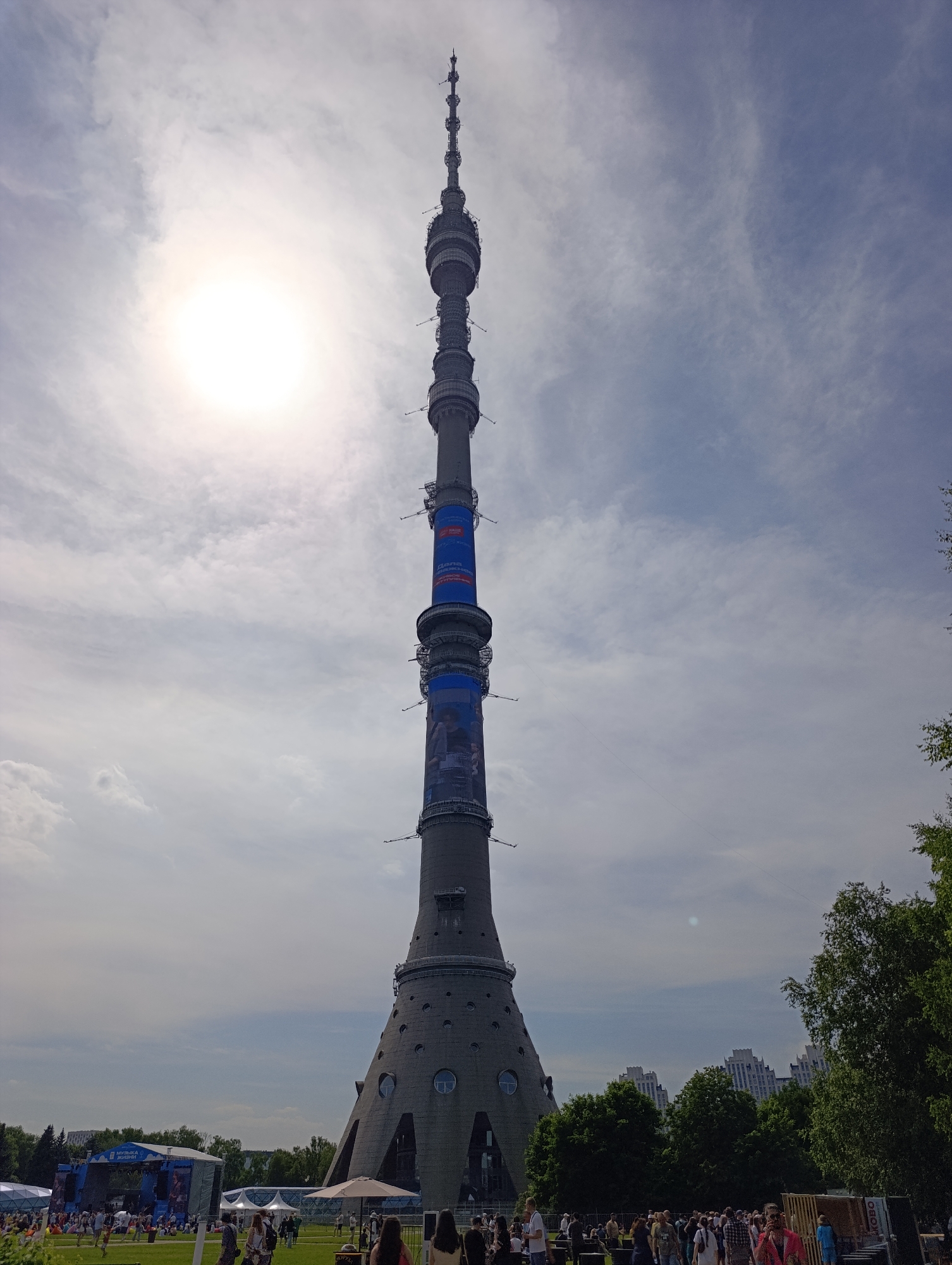 The image presents the Ostankino Tower in Moscow, Russia, captured from a low angle, emphasizing its height against a bright, partly cloudy sky. The sun, appearing as an overexposed white disc, is positioned in the upper left quadrant. The tower itself is a tall, slender structure of gray concrete and metal, featuring multiple antenna arrays and observation decks. Two prominent blue banners are wrapped around its central sections. The upper banner displays partially visible Cyrillic text, including "ДЕЛАЕМ" (DELAEM - "WE ARE DOING/MAKING"), while the lower banner features an image of a person.
At the base of the tower, a large open-air event is underway on a grassy field. To the left, a stage with a blue backdrop is set up, emblazoned with the white Cyrillic text "МУЗЫКА ЖИЗНИ" (MUZYKA ZHIZNI - "MUSIC OF LIFE"). A substantial crowd of people, dressed in casual attire, occupies the foreground, some walking and others standing or observing the event. Several white temporary structures, possibly tents or canopies, are scattered across the field, alongside a white patio umbrella. Lush green trees and foliage line the periphery of the scene on both the left and right sides. In the background, above the treeline, distinctive white multi-story buildings with architectural details characteristic of Moscow are visible under a hazy sky.