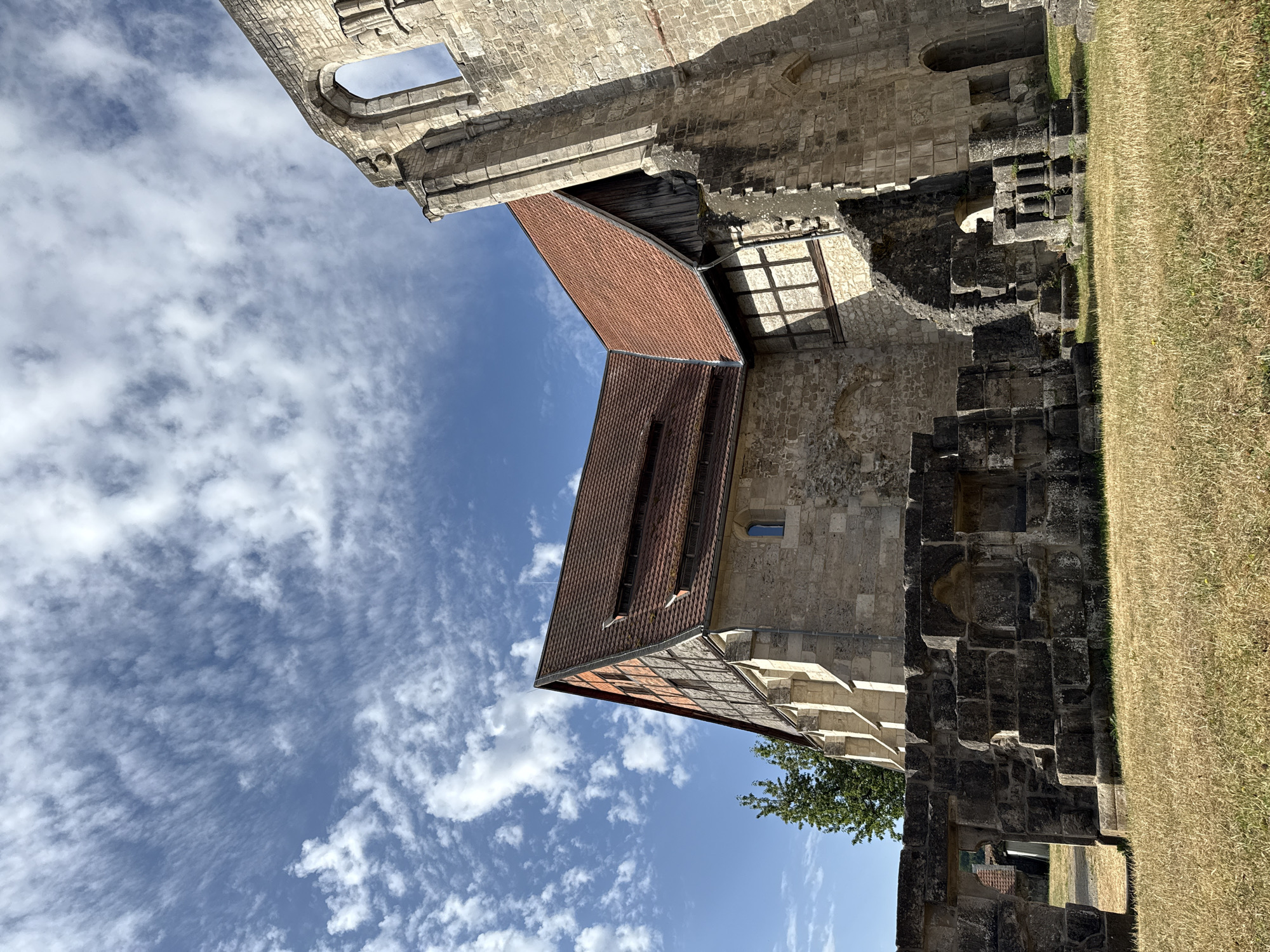 The image shows the ruins of Walkenried Abbey in Walkenried, Germany. A partially reconstructed building with a red tile roof stands next to a crumbling stone structure, suggesting a contrast between preservation and decay. The sky is blue with scattered white clouds. In the foreground, dry grass covers the ground. The stone ruins exhibit architectural elements such as arches and weathered blocks. The scene captures a historical site under a bright sky.