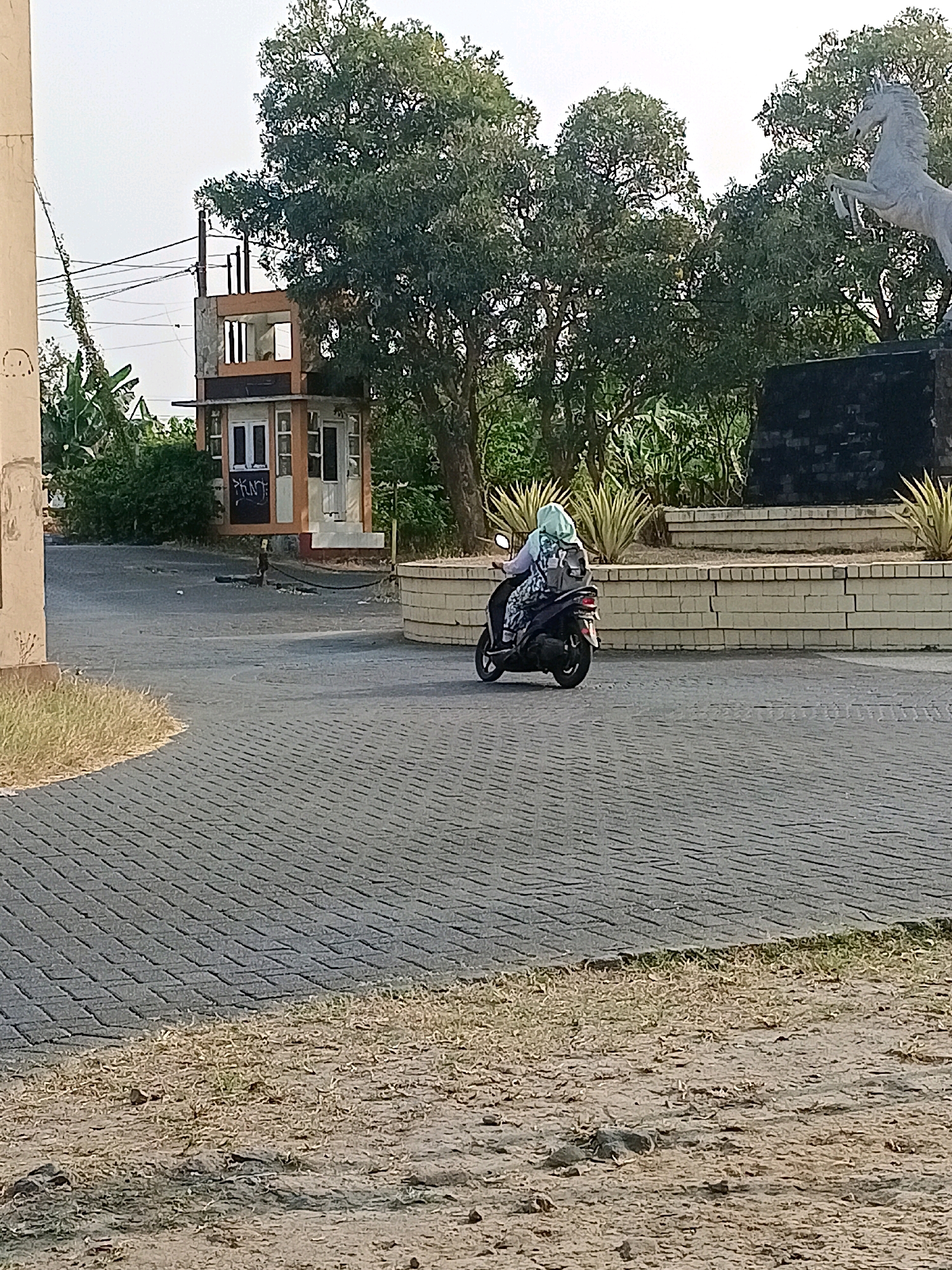 The image depicts a scene in Tawangsari, Indonesia, featuring a woman riding a scooter on a paved road. The scooter is dark-colored and appears to be in motion, with the woman wearing a light blue hijab and a patterned outfit, along with a backpack. In the background, a multi-story building with an open structure and windows is visible, possibly a guardhouse or small office. Green trees and foliage surround the area, and a large stone sculpture of a horse is present on a raised platform to the right. The road is constructed with interlocking gray pavers, and the surrounding ground is a mix of dry grass and dirt. The sky is overcast, indicating daylight.