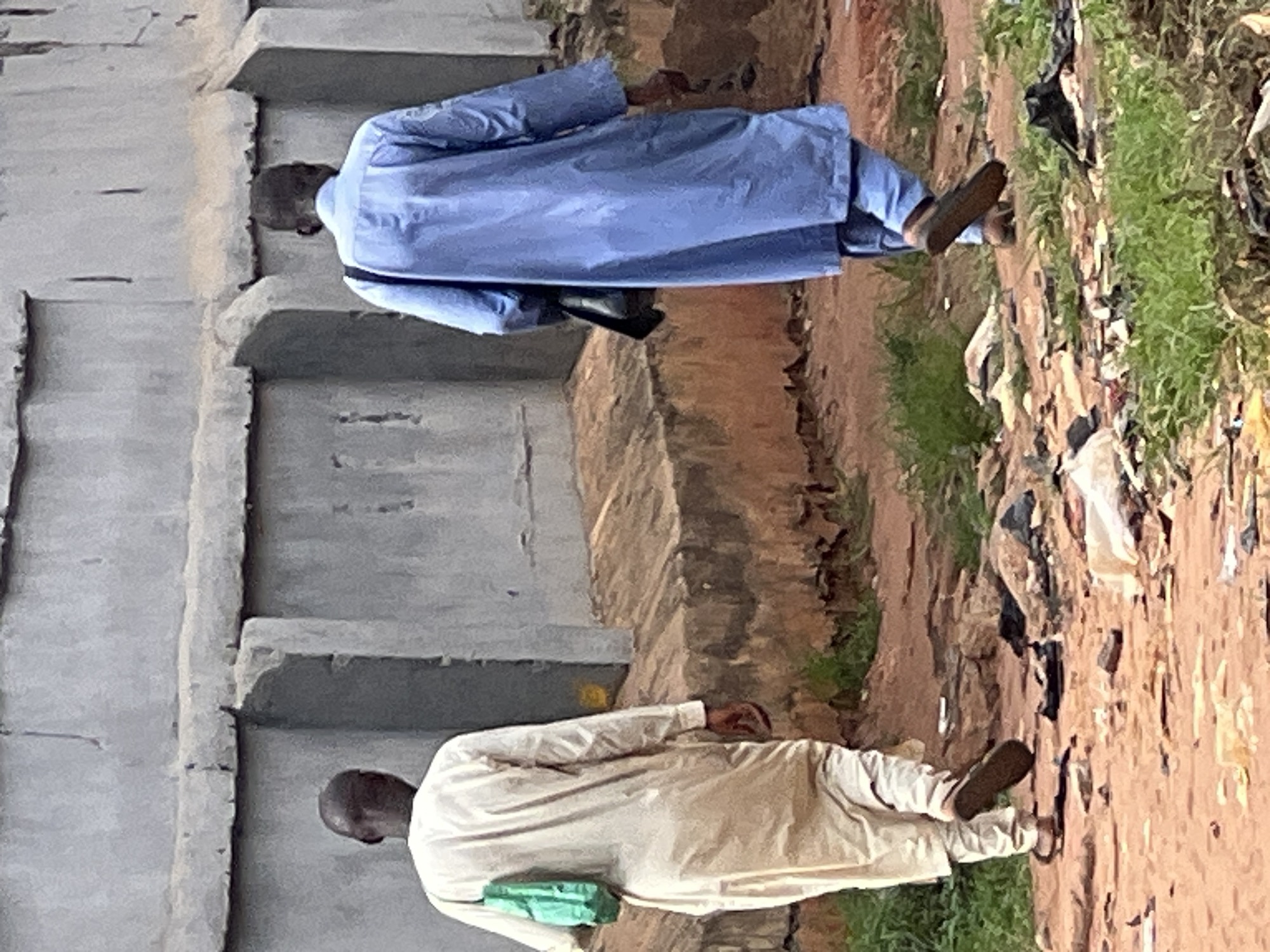 Two men are walking on a dirt road in Obosi, Nigeria, passing a grey concrete structure on the left and trash and vegetation on the right. The man on the top of the frame is wearing a light blue robe and the man on the bottom is wearing a light beige robe. Both men are wearing dark sandals. The road surface is uneven and littered with debris.