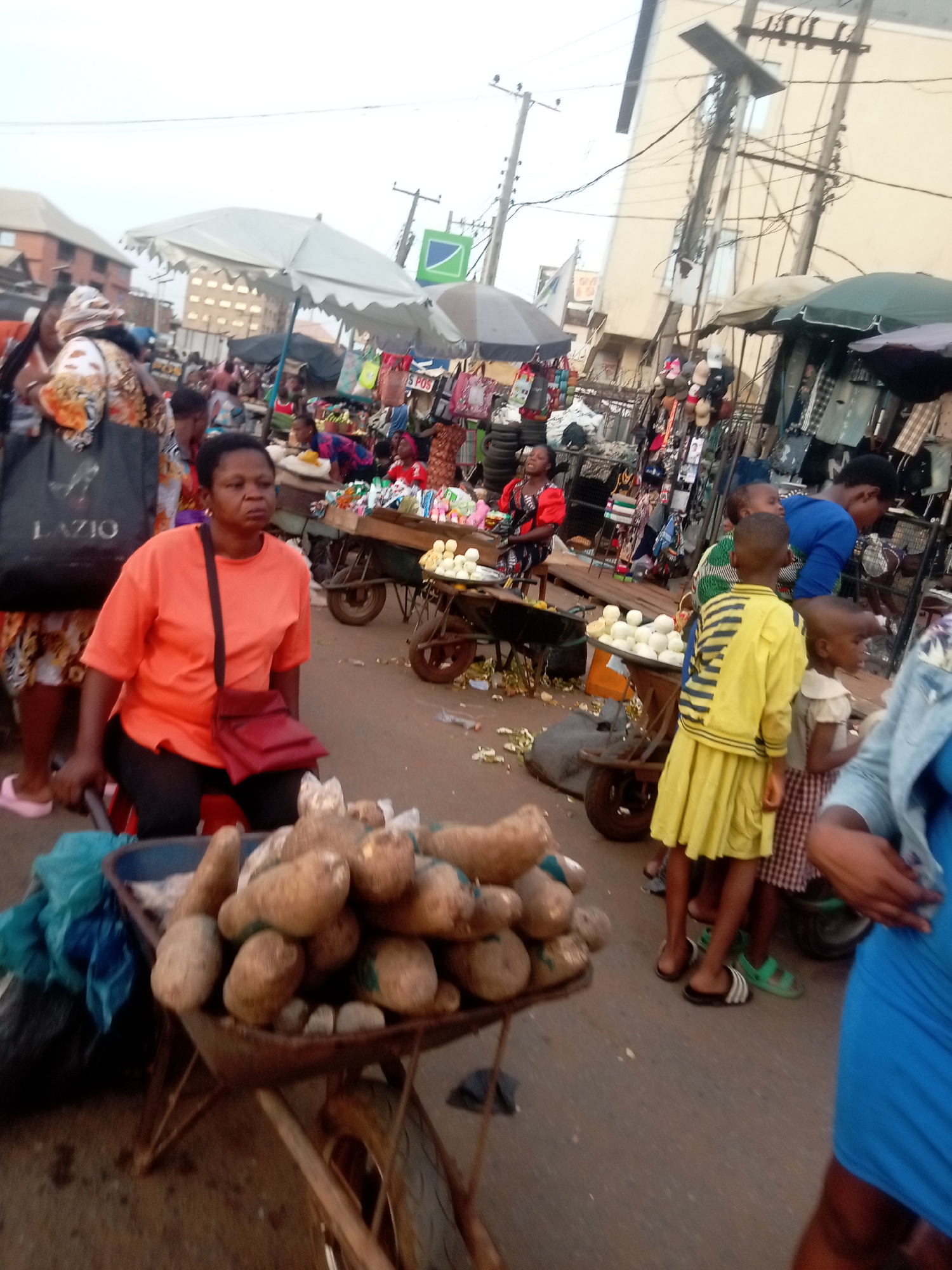 The image depicts a busy street scene, likely a marketplace, in Nkpor, Nigeria. A woman wearing an orange t-shirt and black pants sits on a red stool next to a wheelbarrow filled with yam tubers. The wheelbarrow sits on the street. The woman has a red purse strapped over her shoulder. In the background, a dense crowd of people is visible amidst stalls and vendors displaying various goods under umbrellas. A child wearing a yellow and blue striped shirt and yellow shorts stands near the center of the frame. Other people are visible as well. The street is paved and the environment appears to be bustling with commercial activity. The background shows buildings and overhead power lines typical of an urban setting.