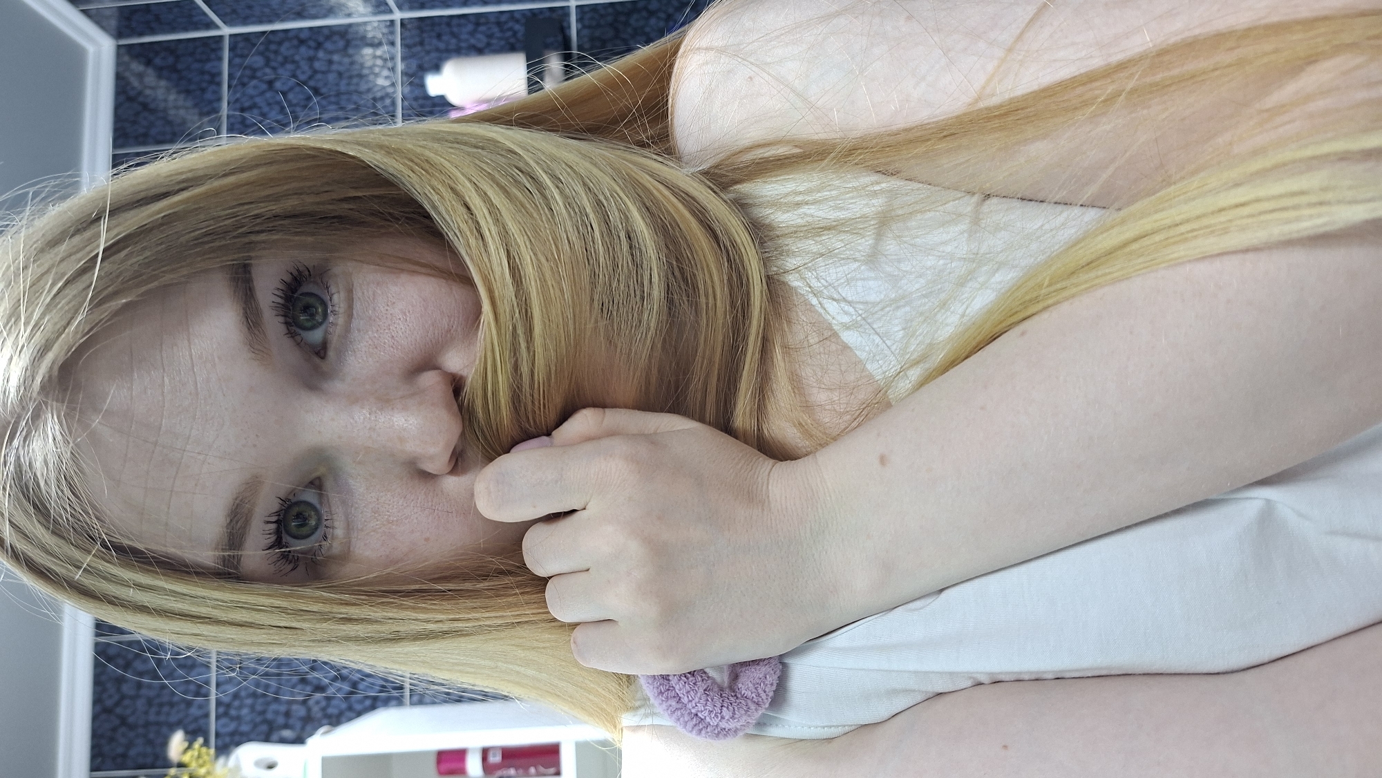 A close-up, tilted, selfie-style image shows a young, fair-skinned woman with light blonde hair and green eyes. She is holding a portion of her hair in front of her mouth. Her face shows freckles. She is wearing a white top. A light purple, knitted item is visible on the lower part of the shirt. The background appears to be a bathroom with blue and white tile, some cleaning products, and a dispenser of some sort. It's impossible to confirm the location within Chaikovsky, Russia, or any specific location without further information.