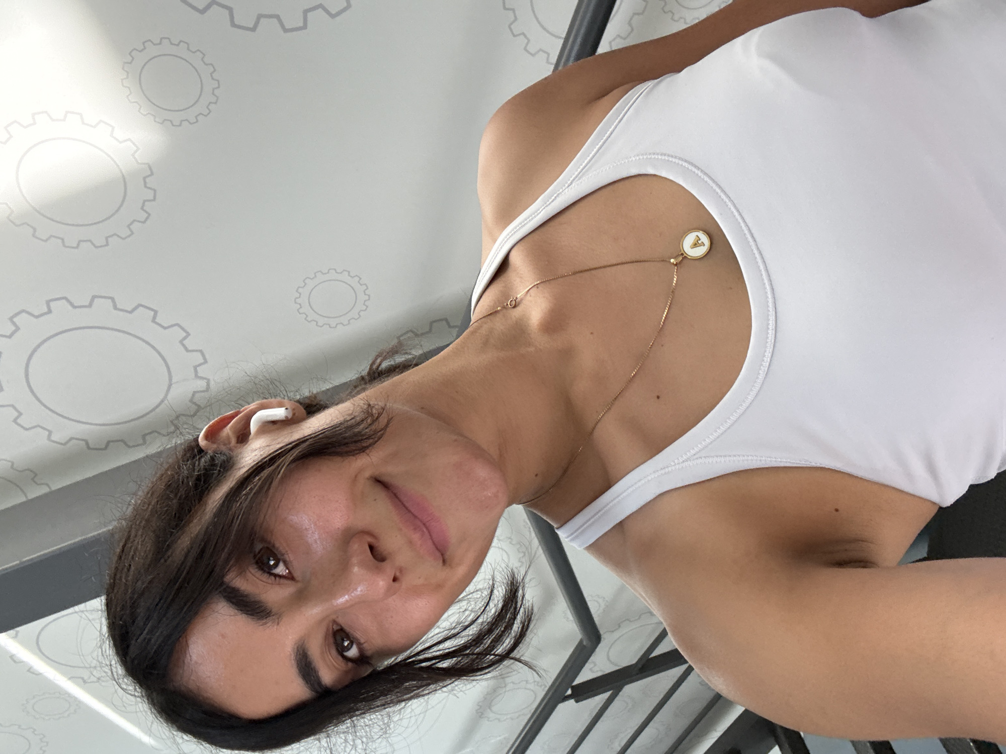 A woman with dark hair, wearing a white tank top and a gold necklace with a white circular pendant, is indoors. She is wearing a white earbud in her left ear. The background features a wall with a gear pattern, and a metallic railing is visible. It is not possible to determine the location within Puebla, Mexico from the image alone.