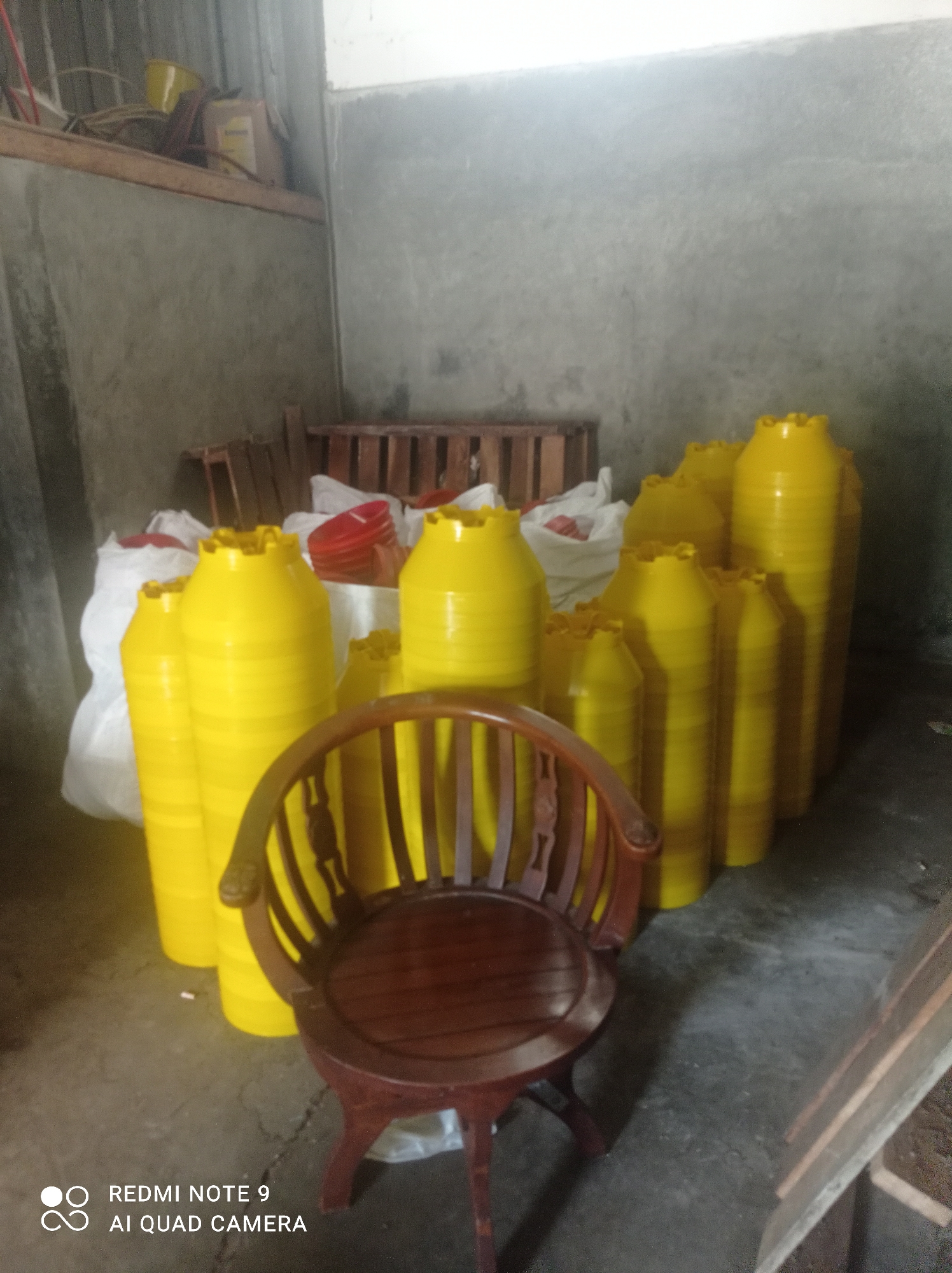 In an indoor setting, possibly a warehouse or storage area, a collection of bright yellow plastic containers are stacked in various sizes. These containers appear to be flower pots or similar vessels, with fluted rims. In the foreground, a dark wooden chair with an ornate back is positioned in front of some of the yellow containers. The floor is concrete, and the walls are a rough, gray concrete. In the background, wooden pallets and white plastic bags are visible, suggesting stored goods. The lighting is diffused, indicating either natural light from an unseen source or artificial lighting. The image includes a watermark at the bottom left corner that reads "REDMI NOTE 9 AI QUAD CAMERA", identifying the device used for capture. The presence of these items suggests a commercial or industrial storage space, potentially located in Cipawon, Indonesia, where such items might be manufactured or stored for distribution.