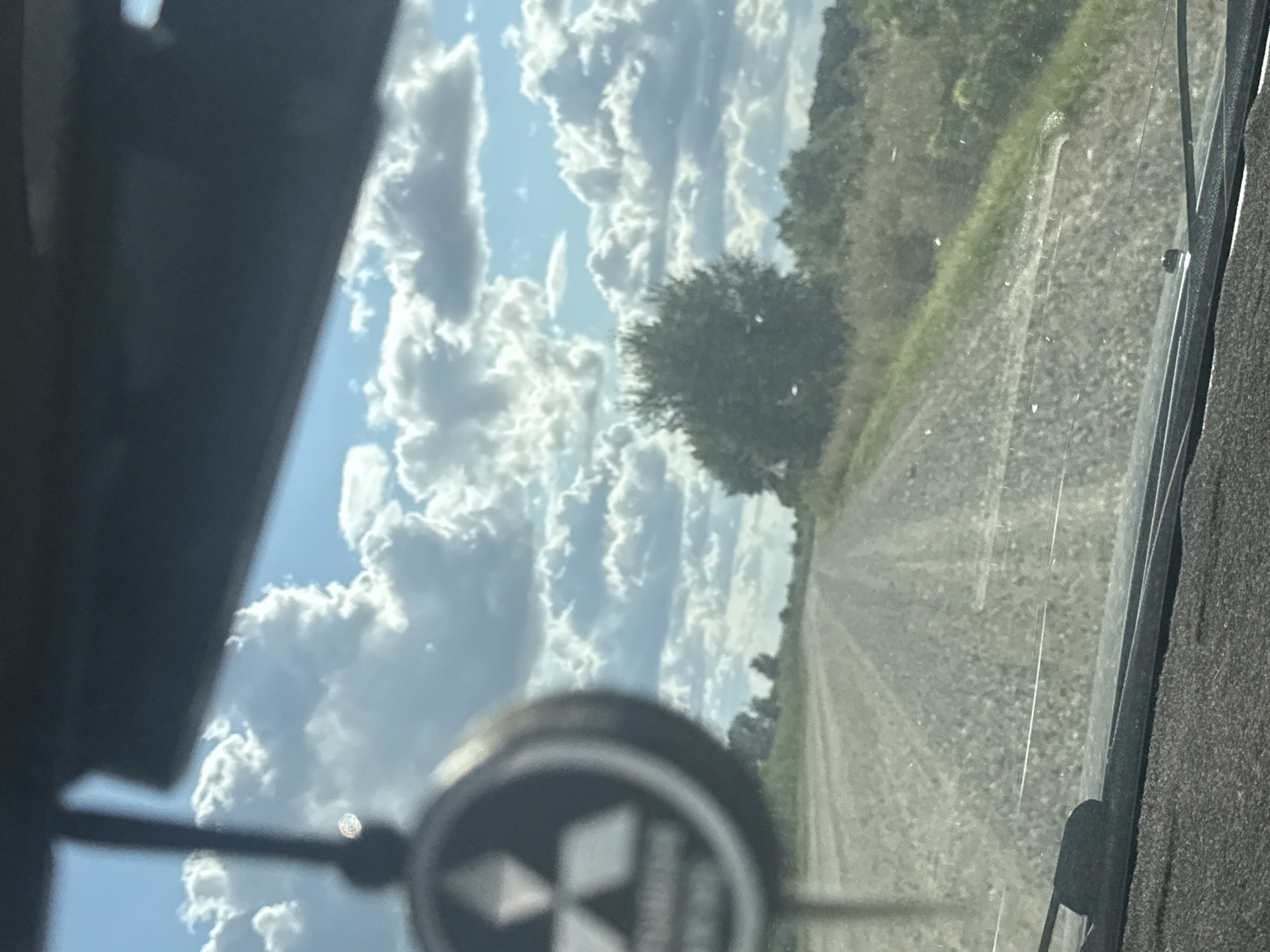The image is a first-person perspective from inside a vehicle, looking out through the windshield. A gravel road stretches into the distance, flanked by dry grass and scattered trees. The sky above is a vibrant blue, filled with large, fluffy white clouds. In the foreground, a dark dashboard and windshield wipers are visible. A small, blurry object hangs from the rearview mirror, featuring a circular emblem with three triangles arranged symmetrically. This scene is not identifiable as being in the city of Васильевка, Russia.